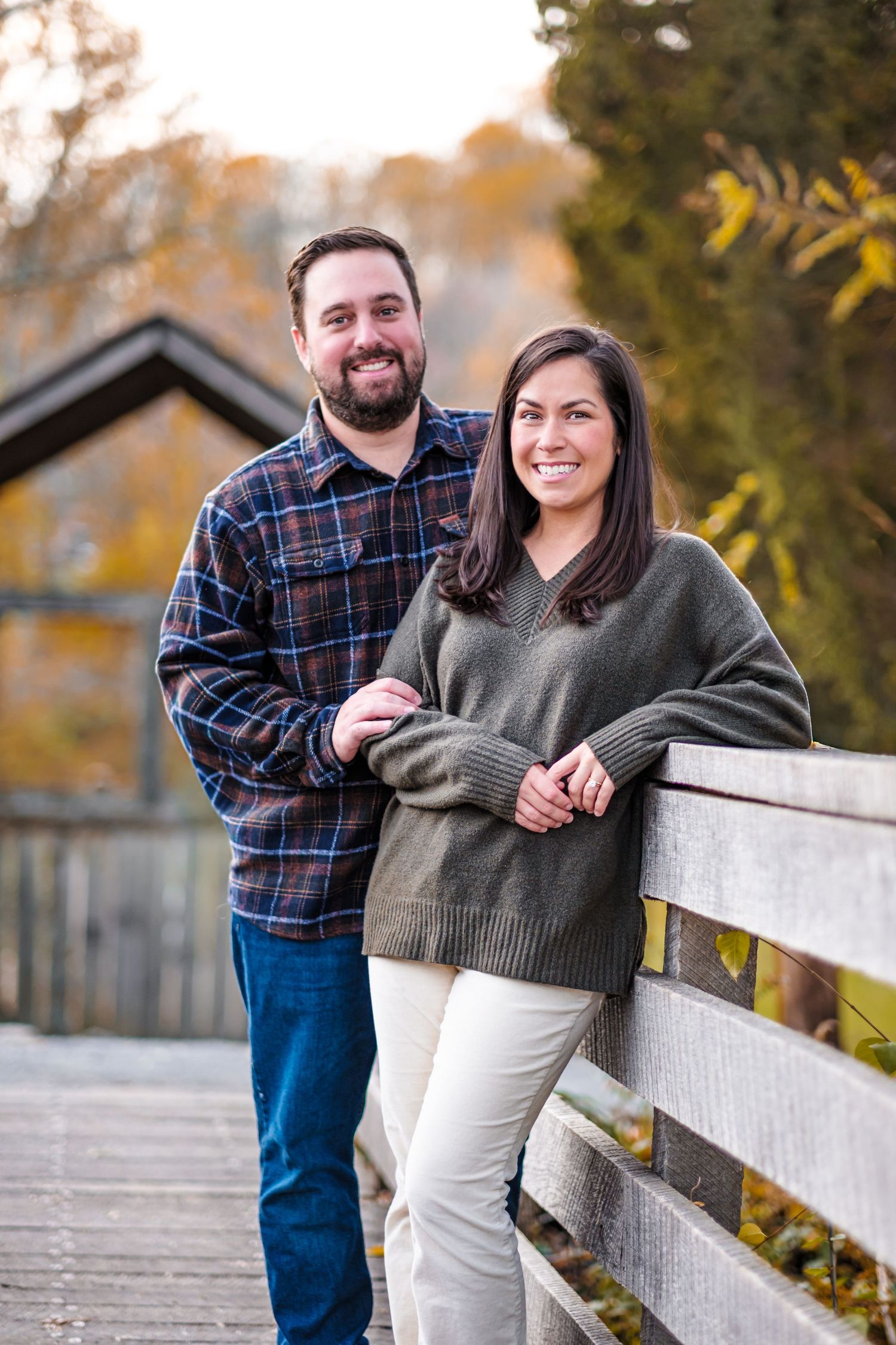 A smiling couple poses on a wooden bridge surrounded by autumn foliage, wearing casual fall attire.