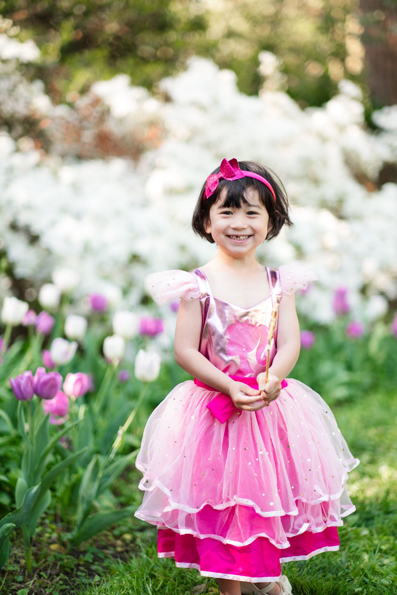Young girl in pink princess dress and fairy wings smiles among tulips and white flowers in garden.