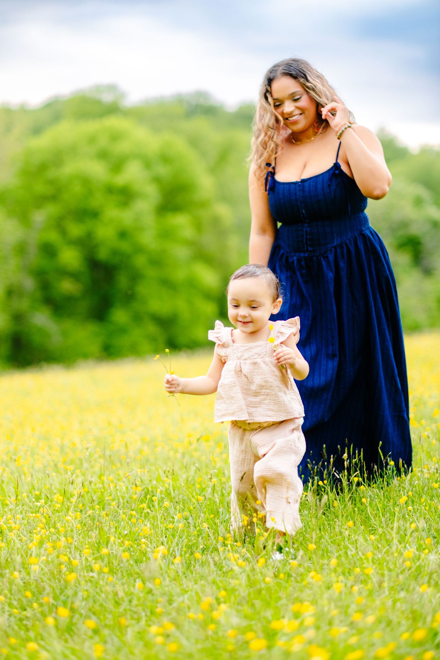 A mother in a blue dress walks with her toddler through a sunny yellow wildflower field with green trees in the background.