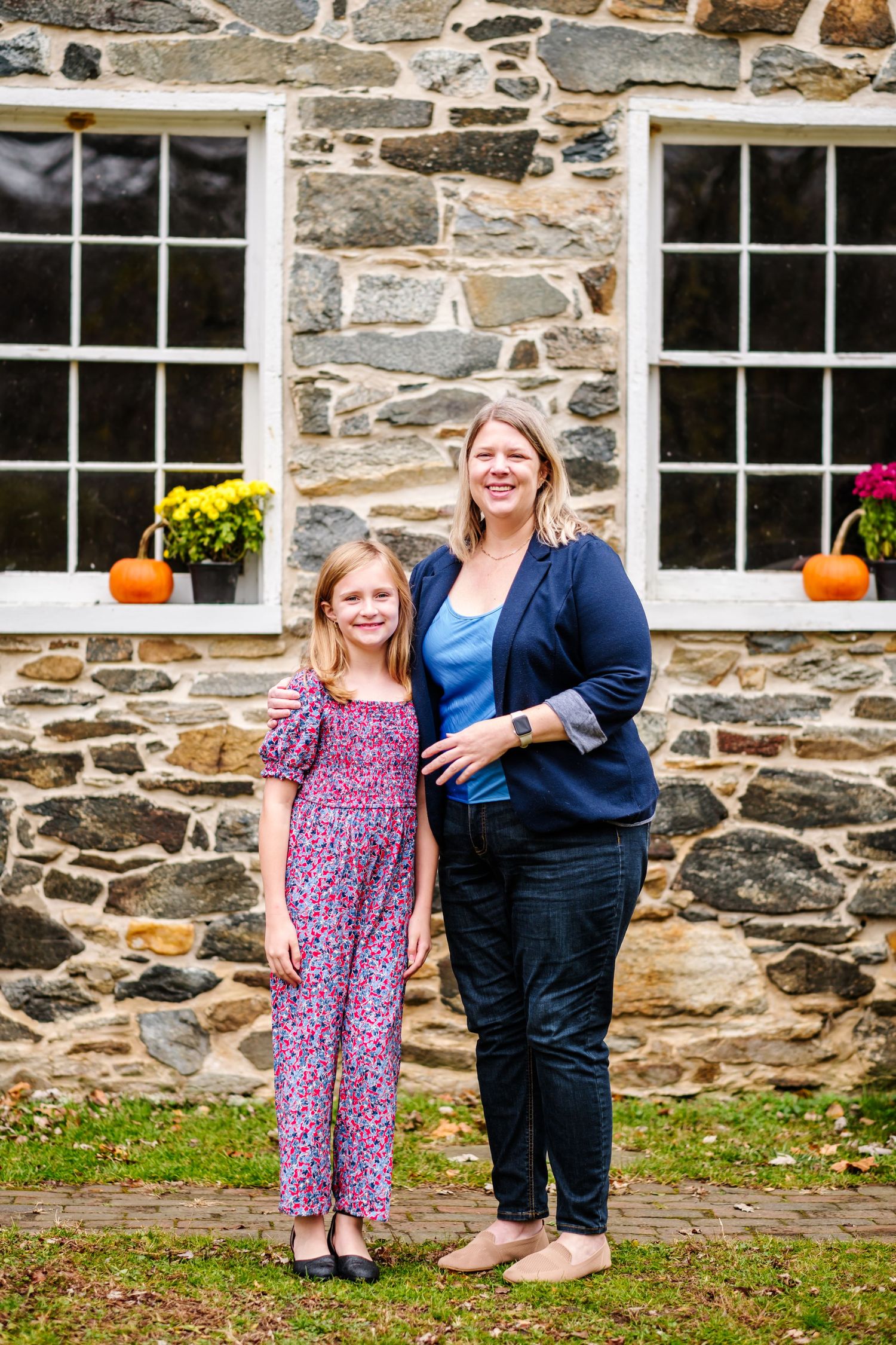 Woman and young girl smiling together in front of a stone building with fall pumpkins and flowers.