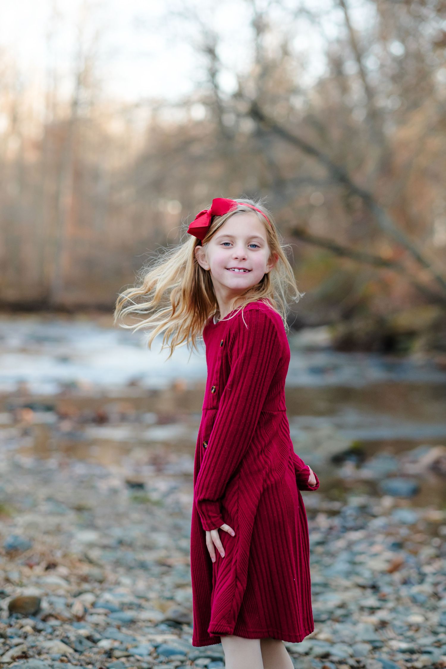 Young girl in red dress with bow smiles by rocky creek, blonde hair flowing in autumn outdoor setting.