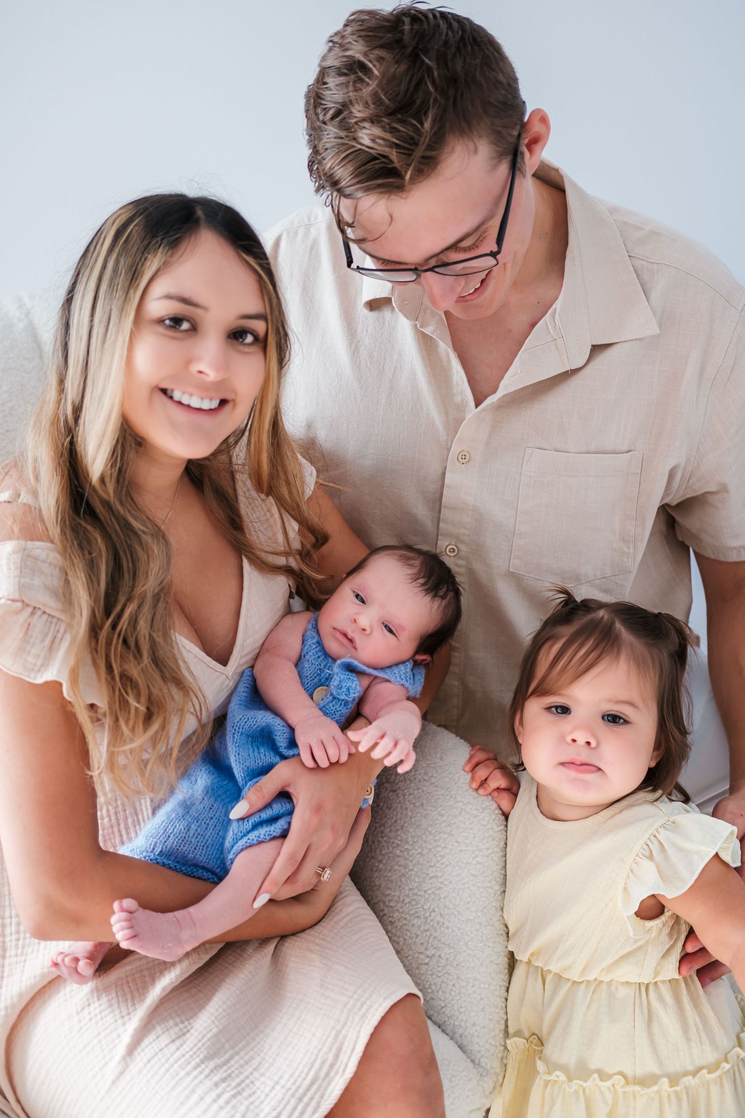 A young family poses together, parents holding a newborn and toddler girl in a bright studio.