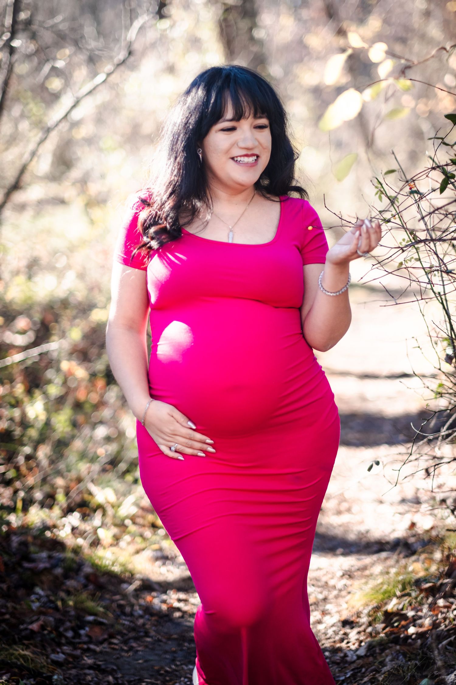 Smiling pregnant woman in pink maxi dress holding her baby bump outdoors in a sunlit forest setting.