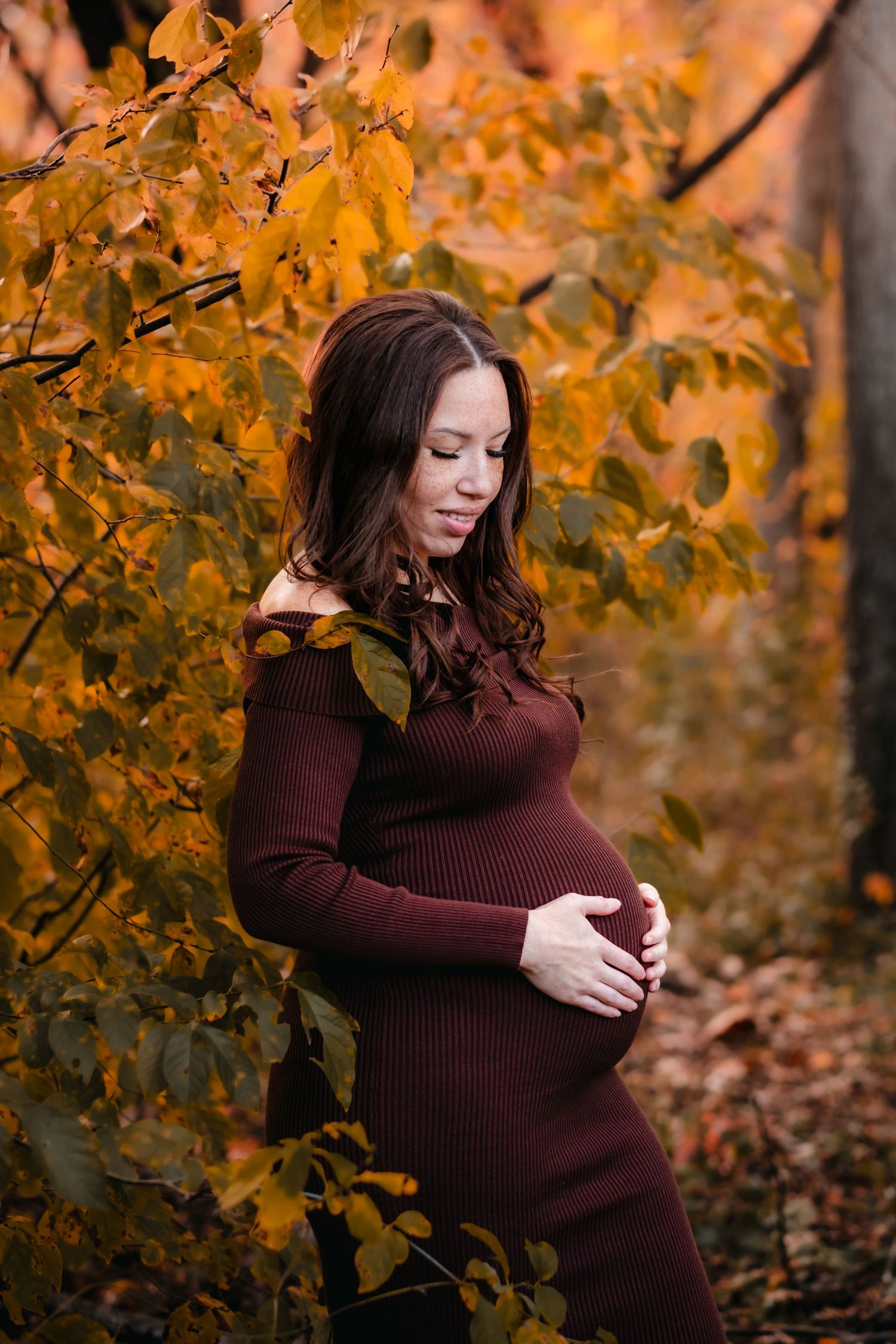 Pregnant woman in burgundy dress cradling baby bump among vibrant autumn foliage in a serene forest setting.