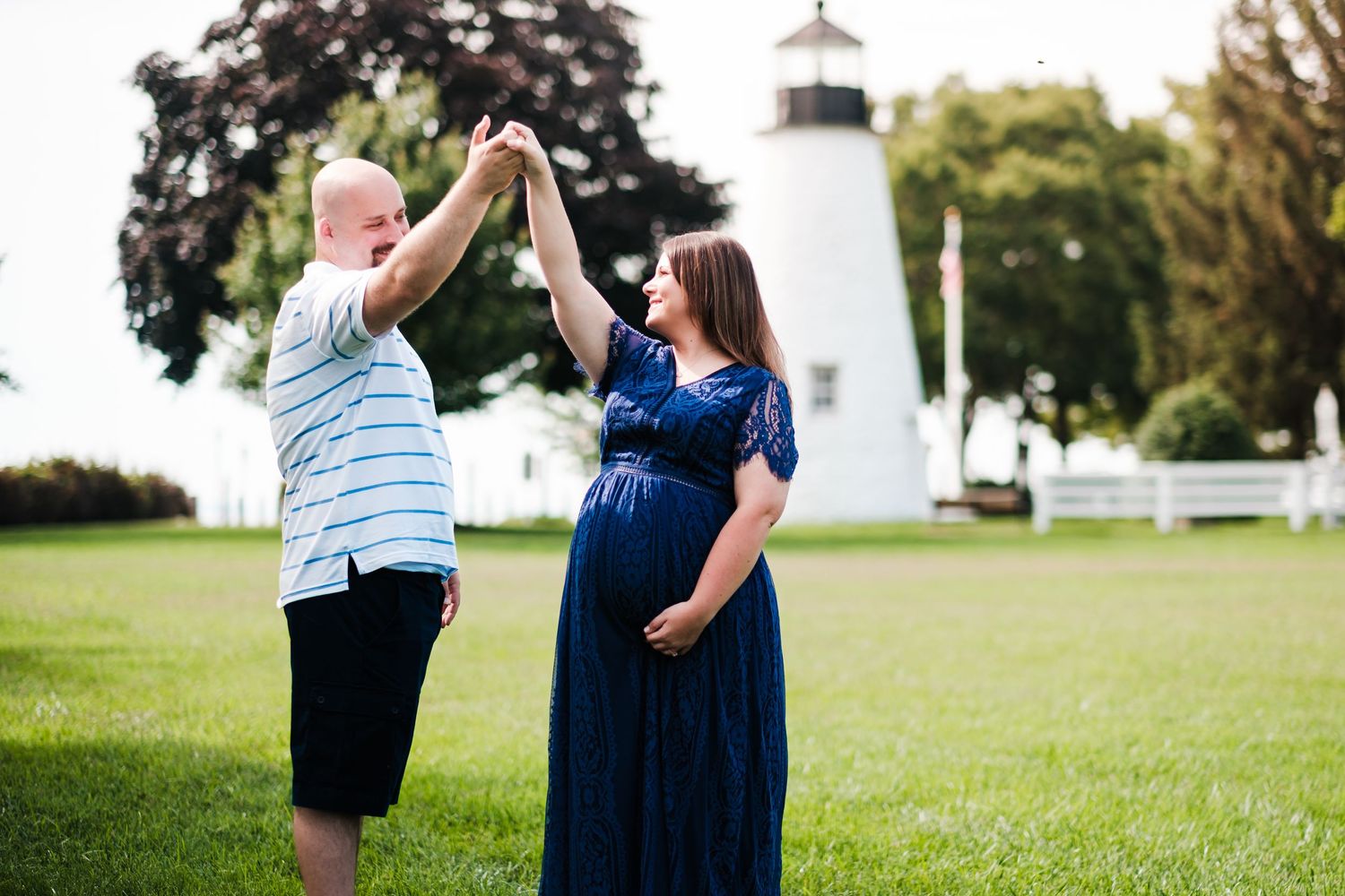 A maternity photo showing two people dancing in front of a white lighthouse on green grass.