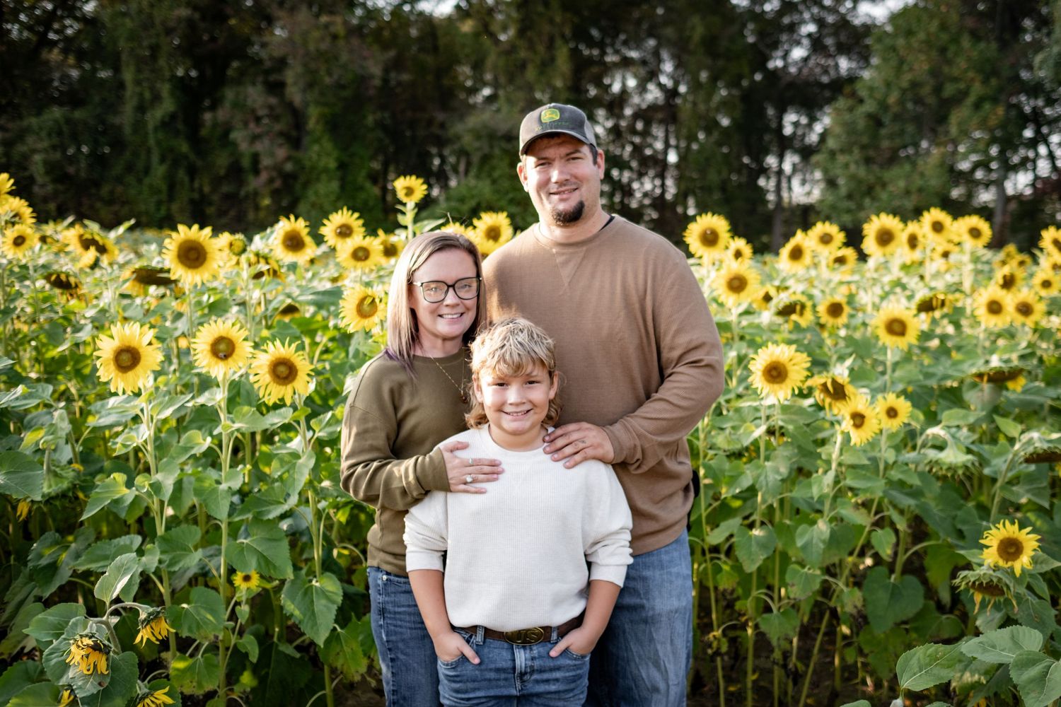 Family of three smiling in a vibrant sunflower field, with tall trees in the background on a sunny day.