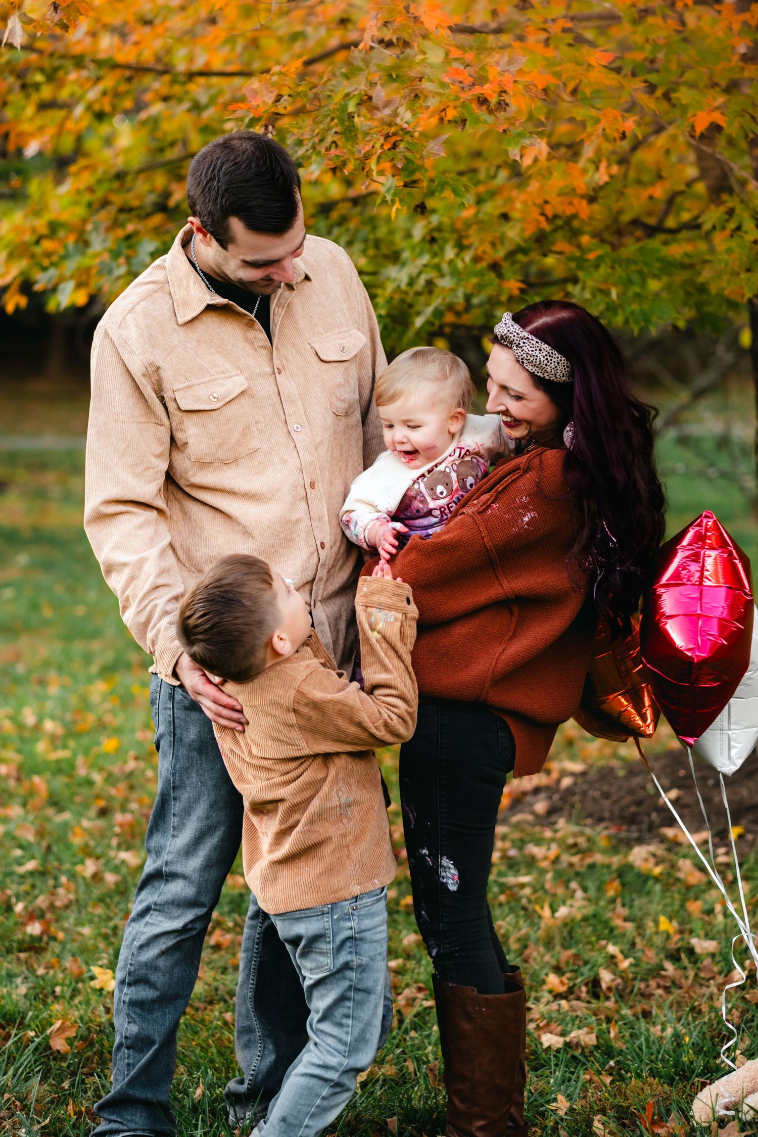 Family of four enjoying autumn outdoors, parents smiling at baby, young boy hugging mom, red balloons, fall foliage background.