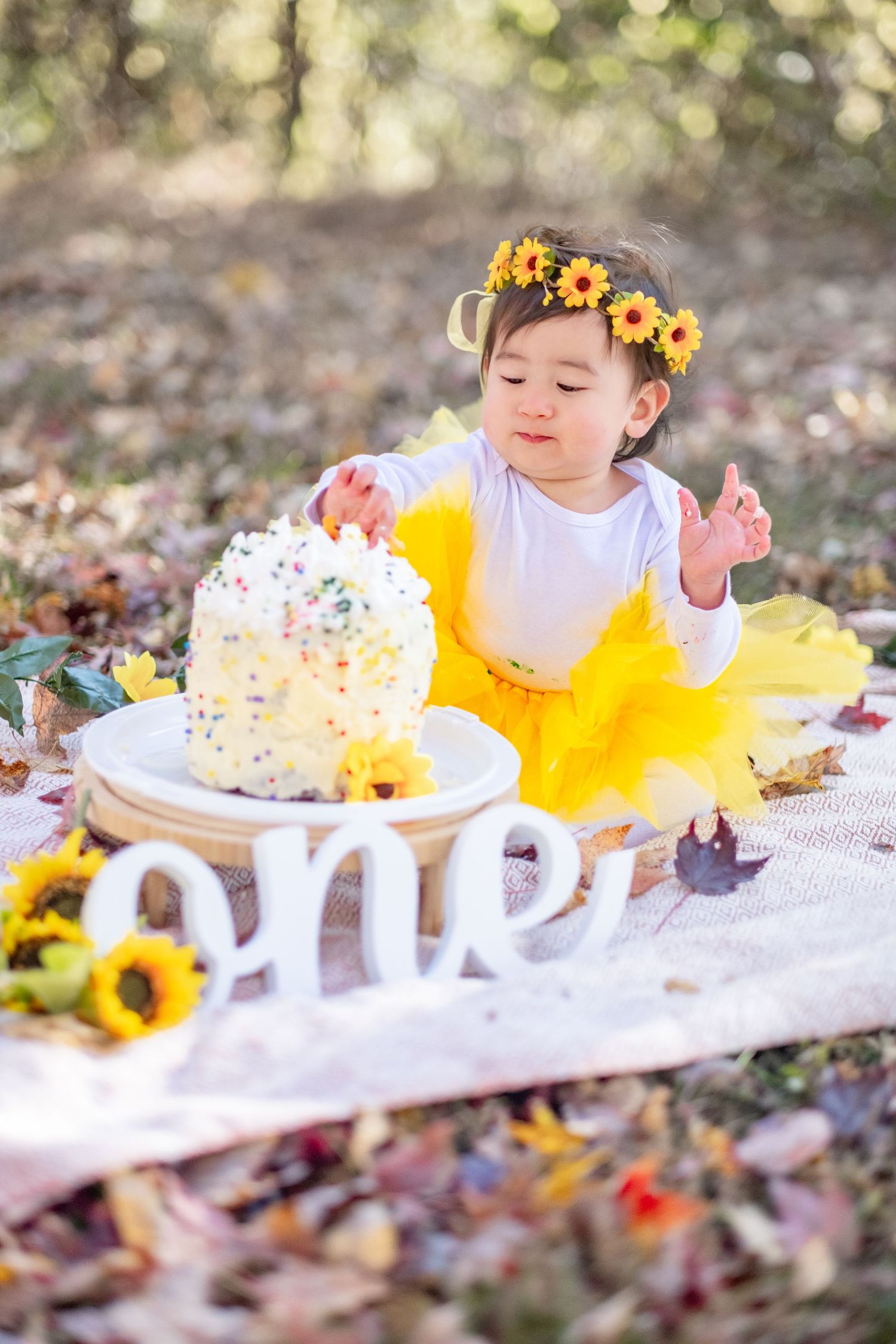 Baby girl in yellow tutu and sunflower crown smashes first birthday cake outdoors with 