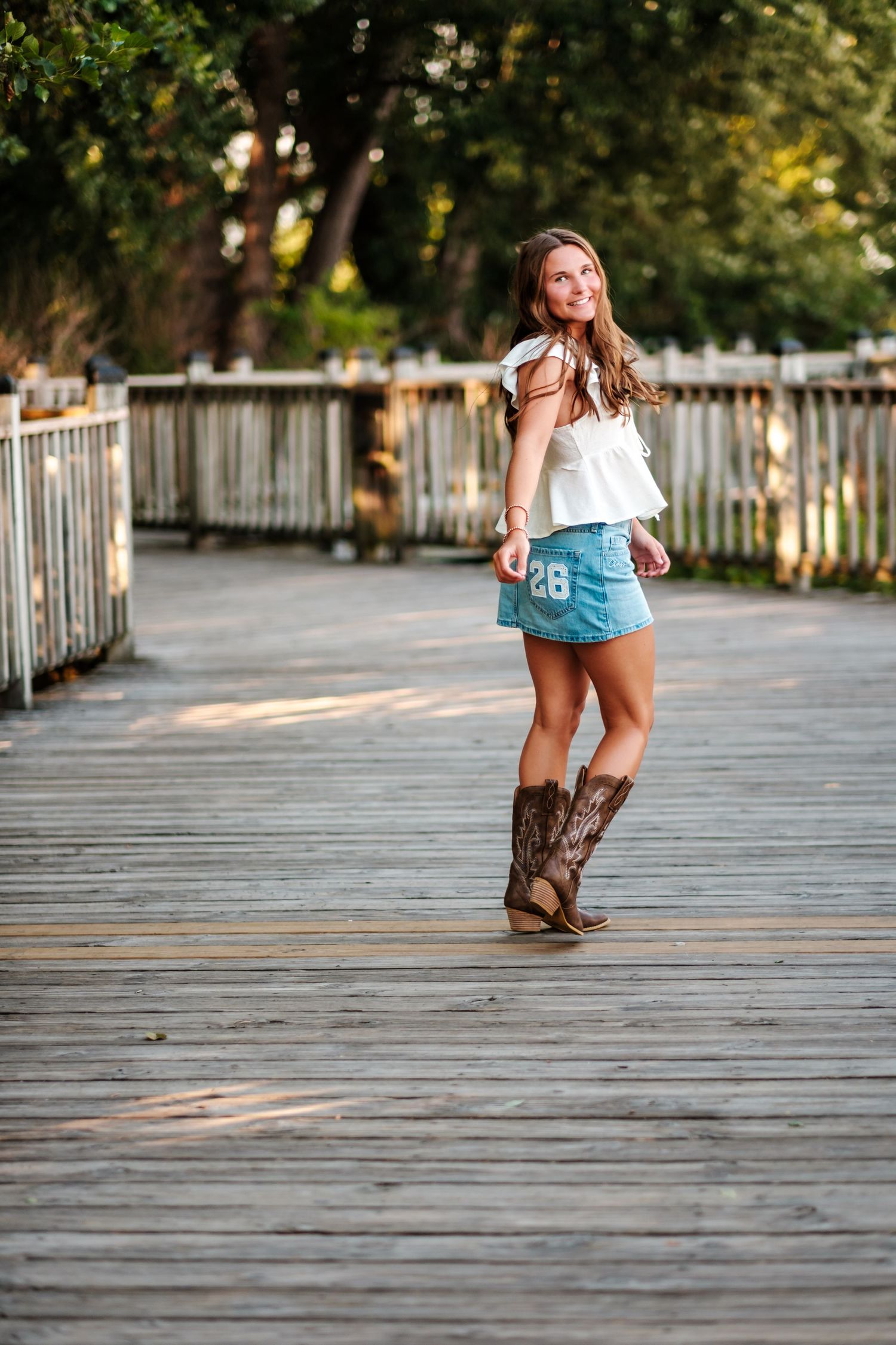 Smiling young woman in white ruffled top, denim mini skirt, and cowboy boots walking on a boardwalk.