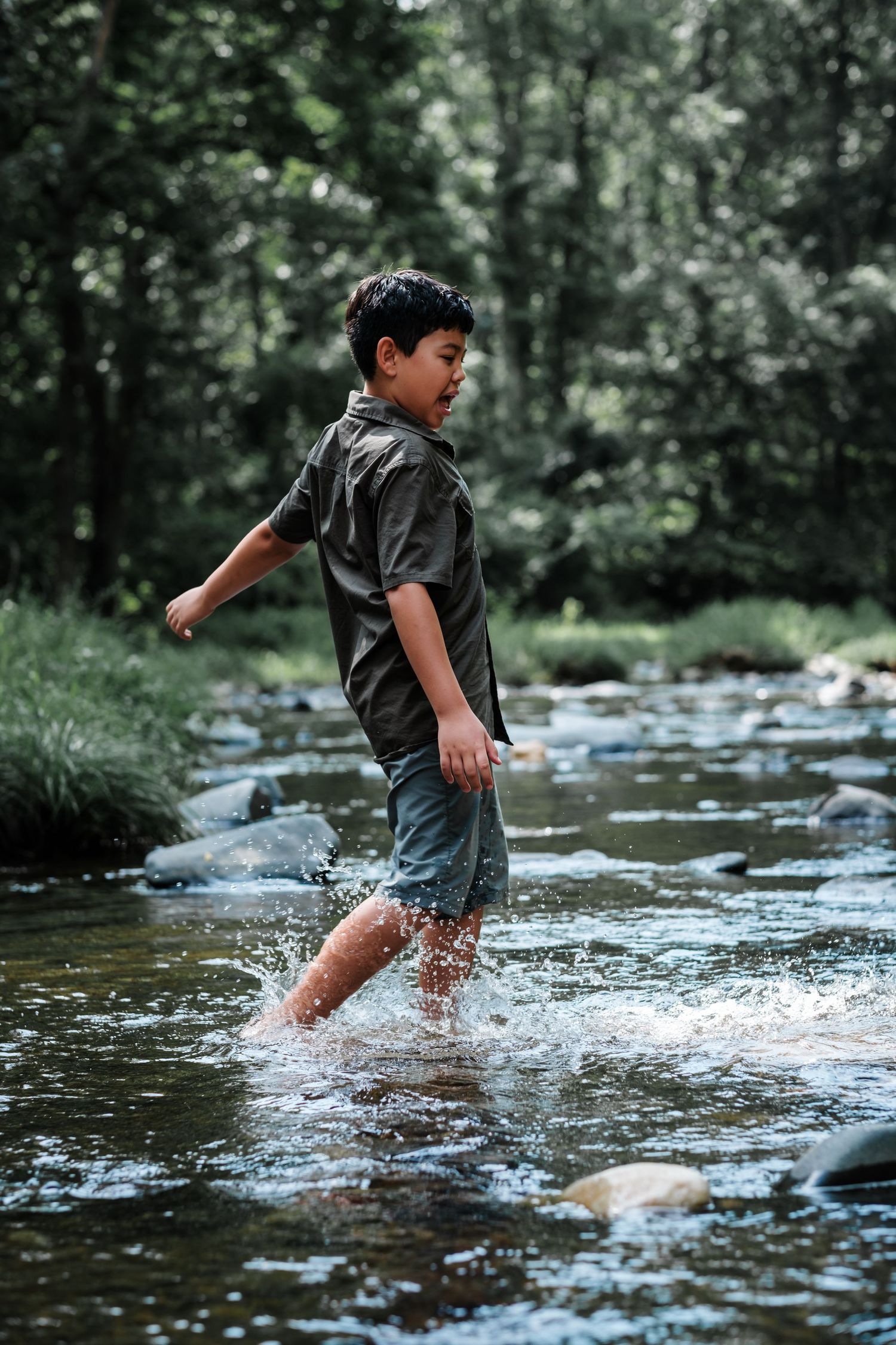 A young boy wades through a shallow rocky stream in a forest, splashing water around him.