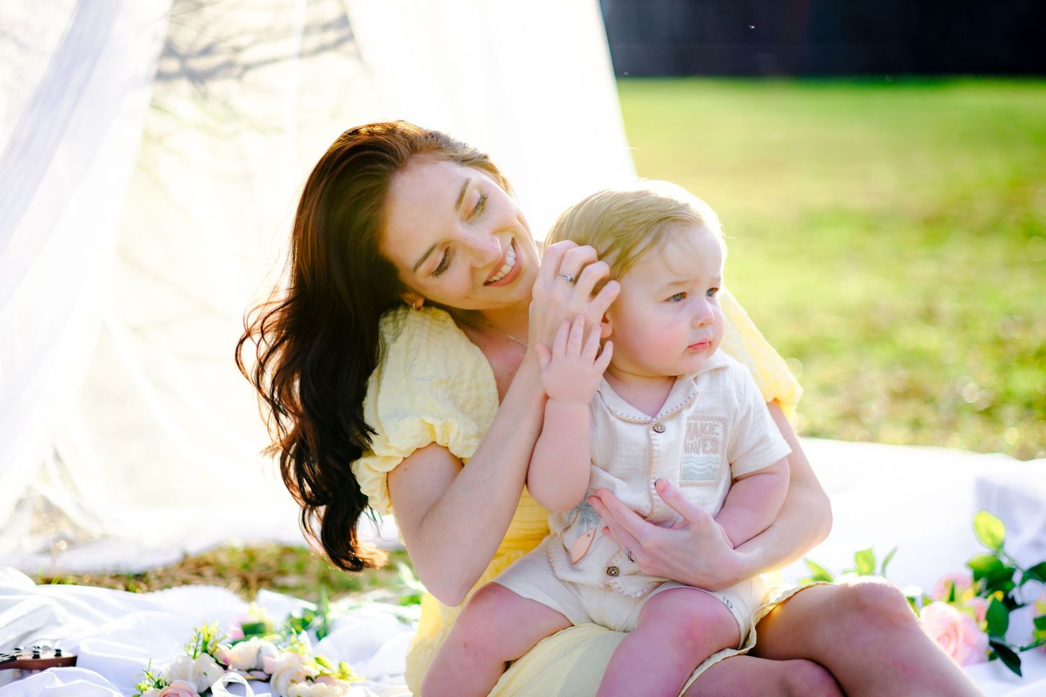 Loving mother in yellow dress tenderly touches baby's face while sitting on blanket in sunny outdoor setting.