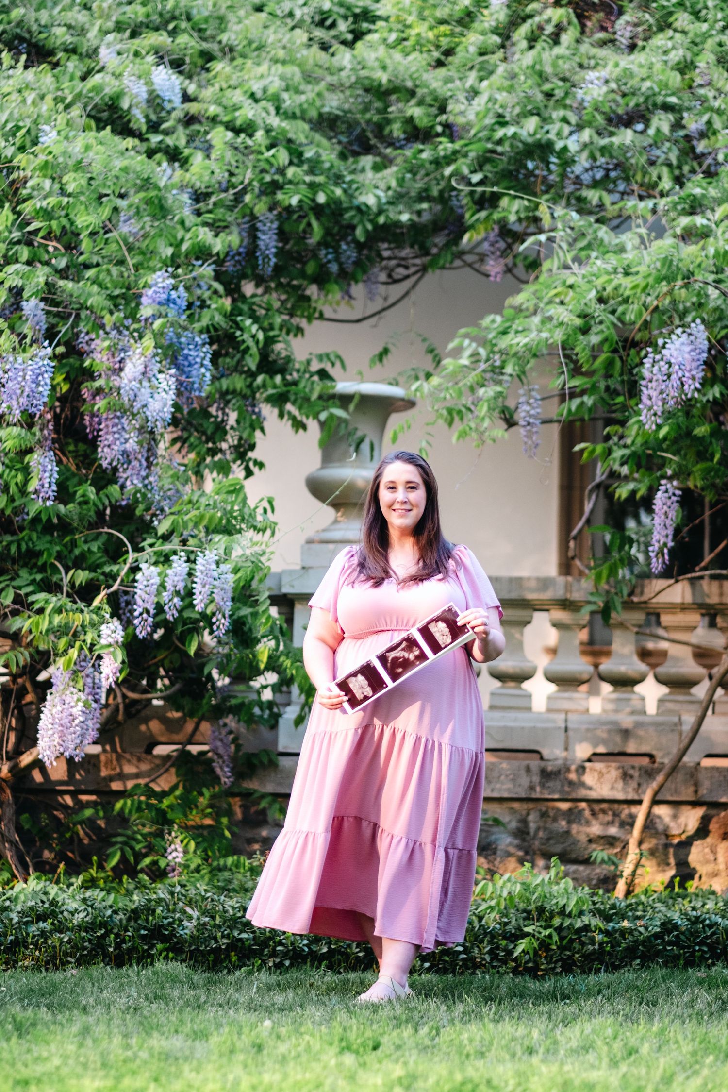 Pregnant woman in pink dress holding ultrasound photos, standing in garden with blooming wisteria.