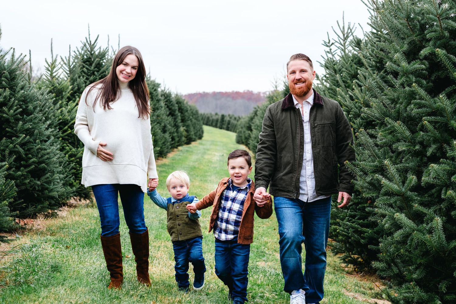 Pregnant woman and man walk with two young boys through Christmas tree farm rows outdoors.