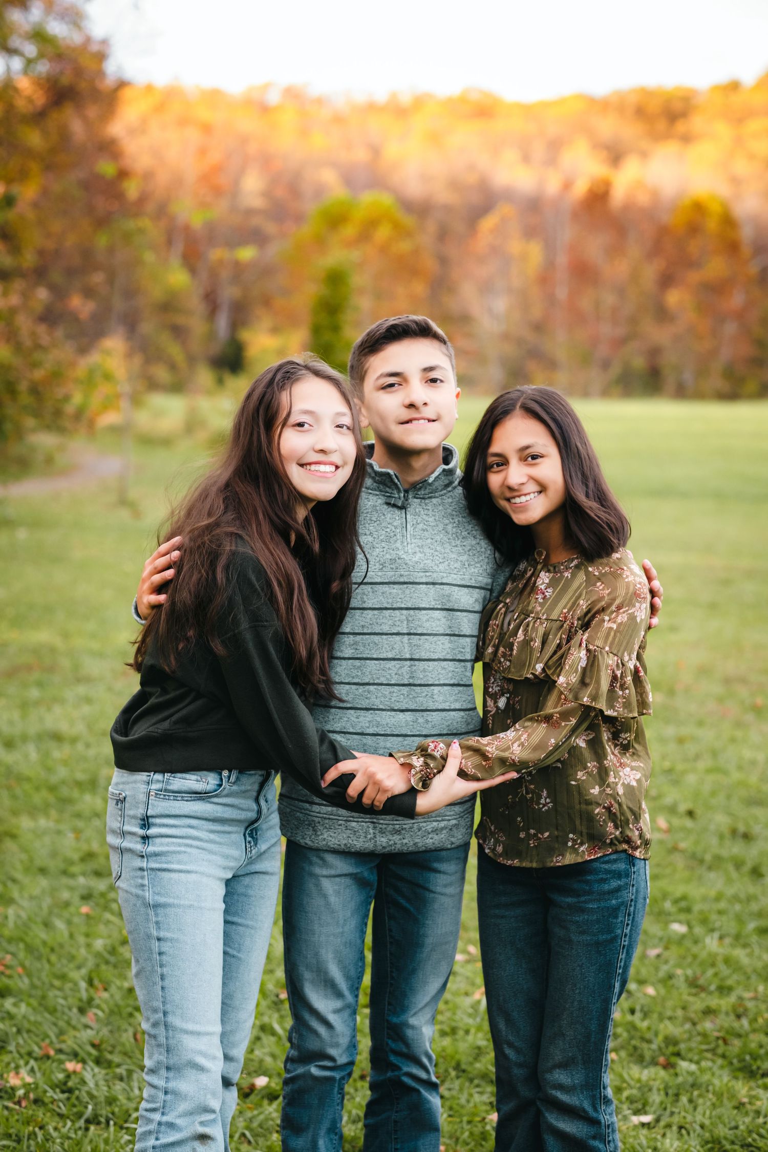 Three smiling teens pose together outdoors in a fall landscape with colorful autumn foliage in the background.
