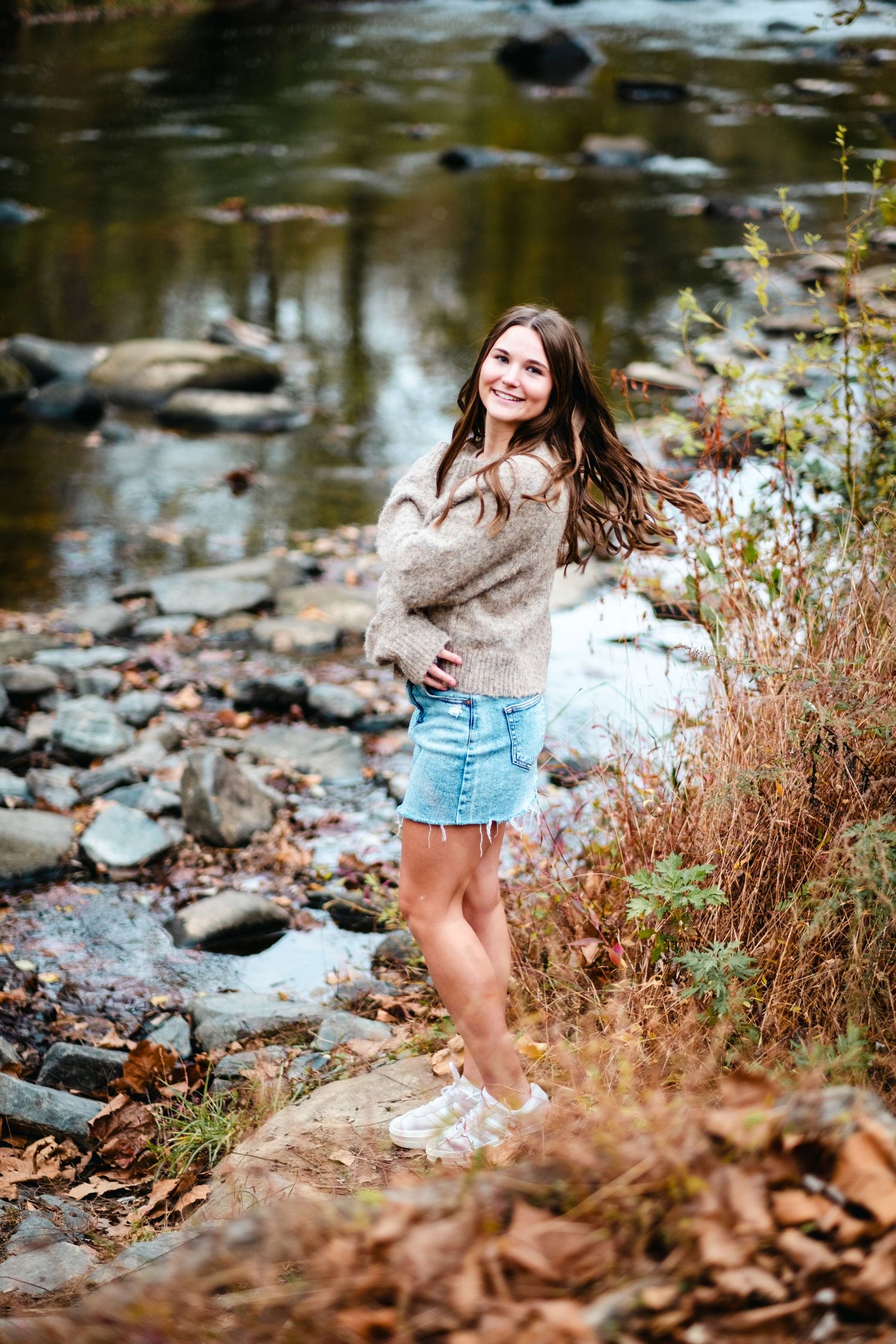 Smiling young woman in cozy sweater and denim skirt poses by a rocky creek surrounded by autumn foliage.