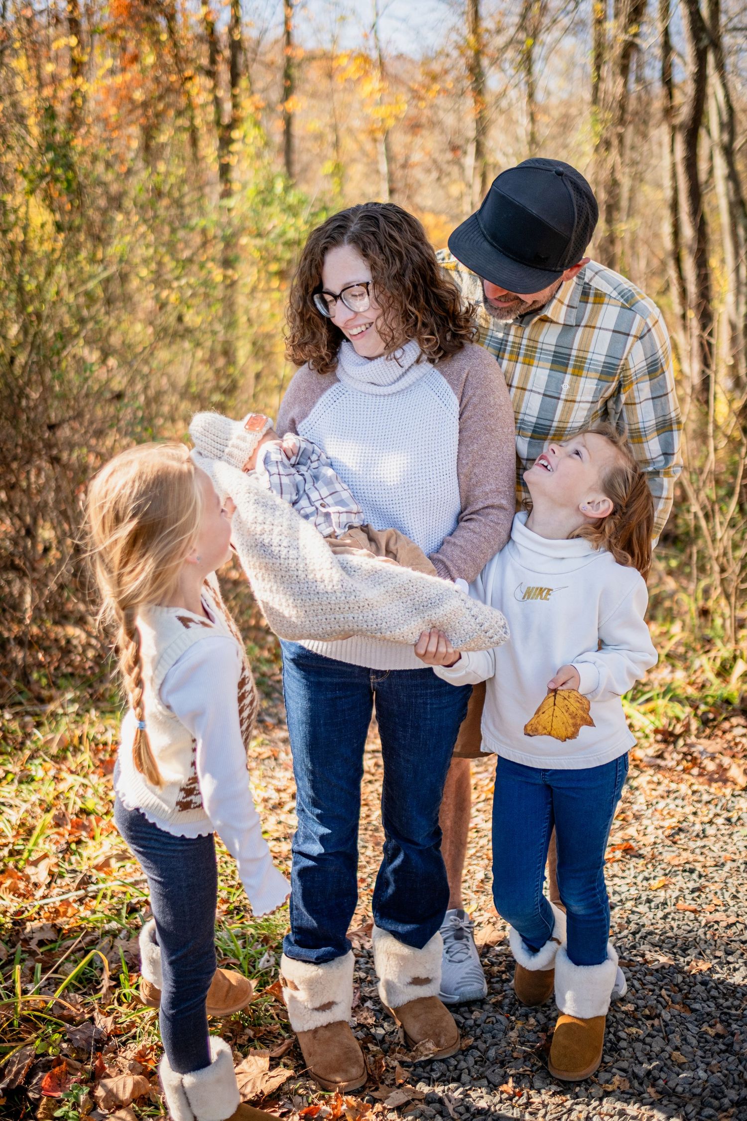 Family of four admiring newborn baby outdoors in autumn forest setting with colorful fall foliage.