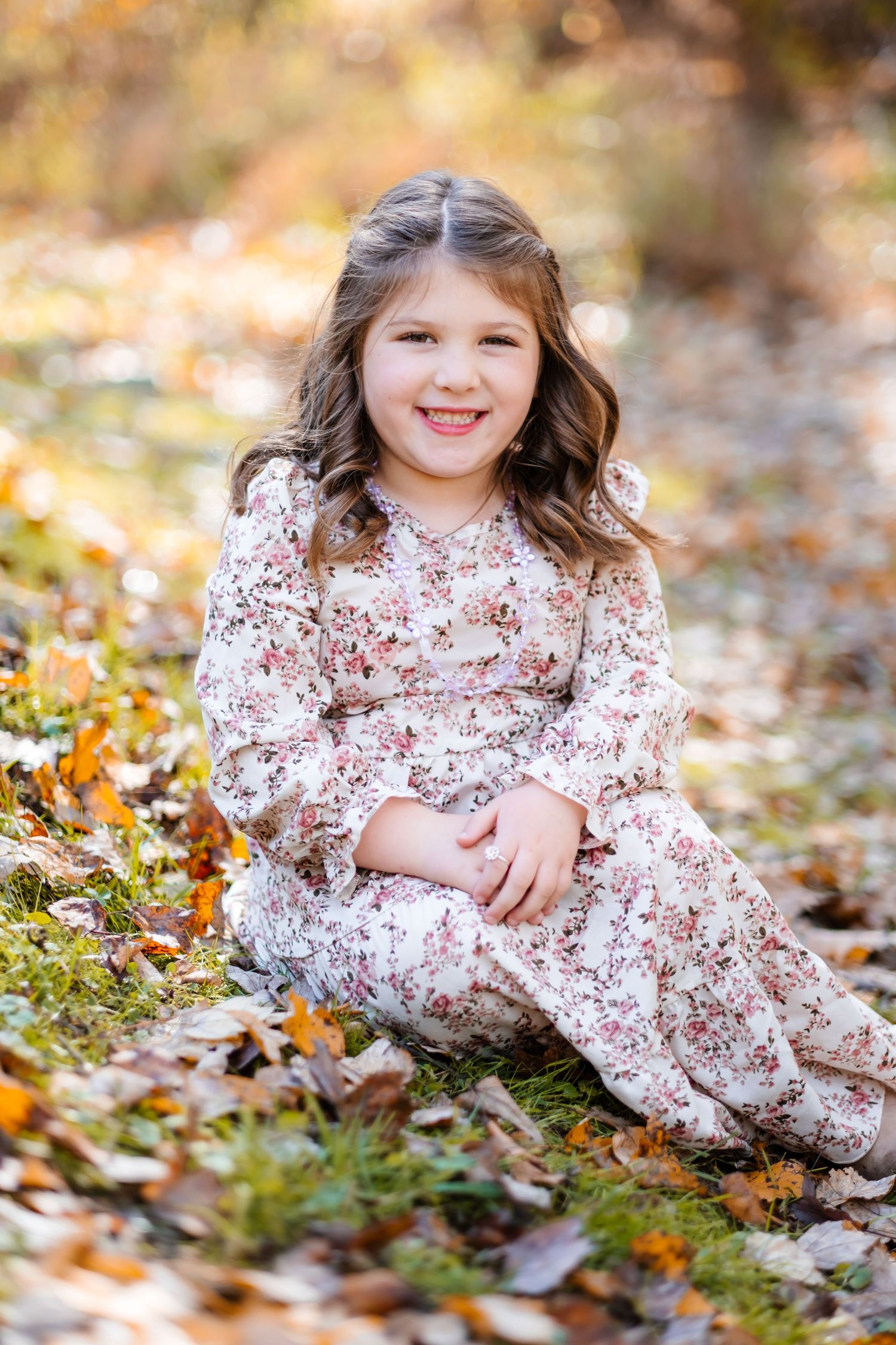 Young girl in floral dress sitting among autumn leaves in a forest, smiling happily at the camera.