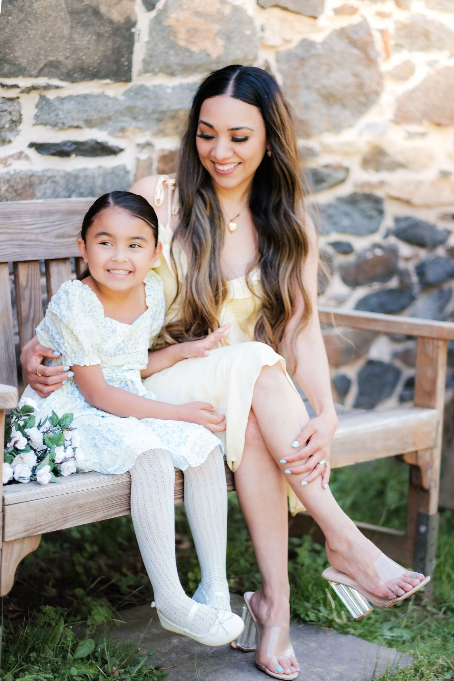 A smiling woman in a yellow dress sits on a wooden bench with a young girl in a white floral dress holding flowers.