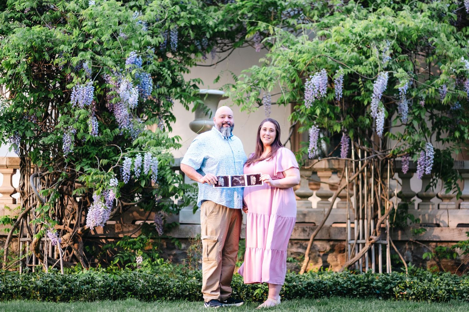 Couple holding ultrasound photos standing under blooming wisteria in a lush garden during maternity photo session.