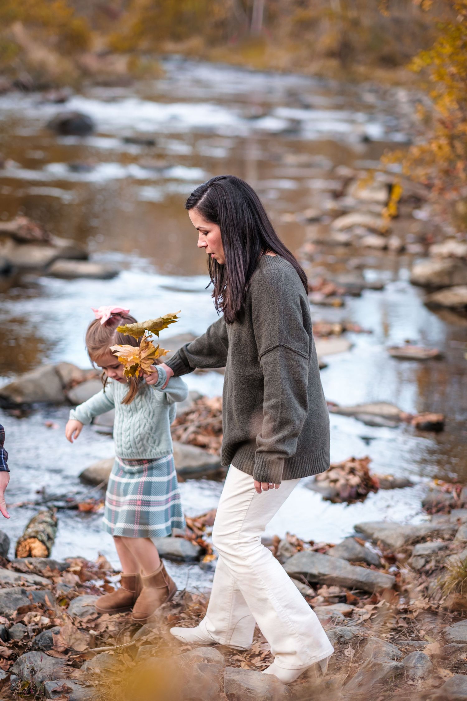 Mother and toddler daughter playing with autumn leaves by a rocky creek in fall.