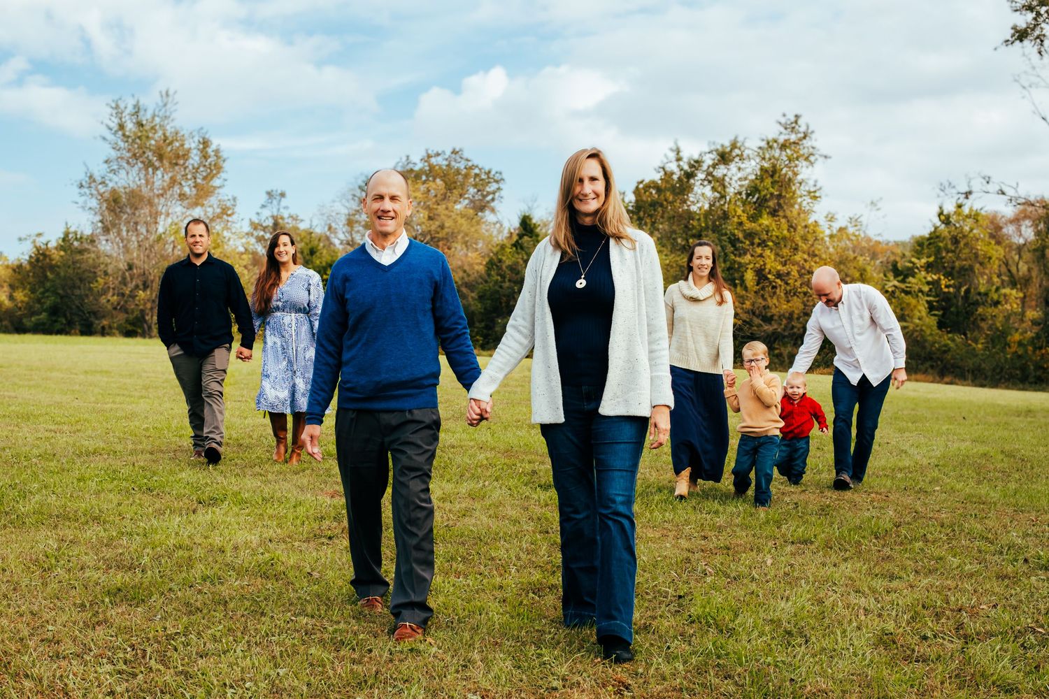 Multi-generational family walking together in a green field during fall, holding hands and smiling outdoors.