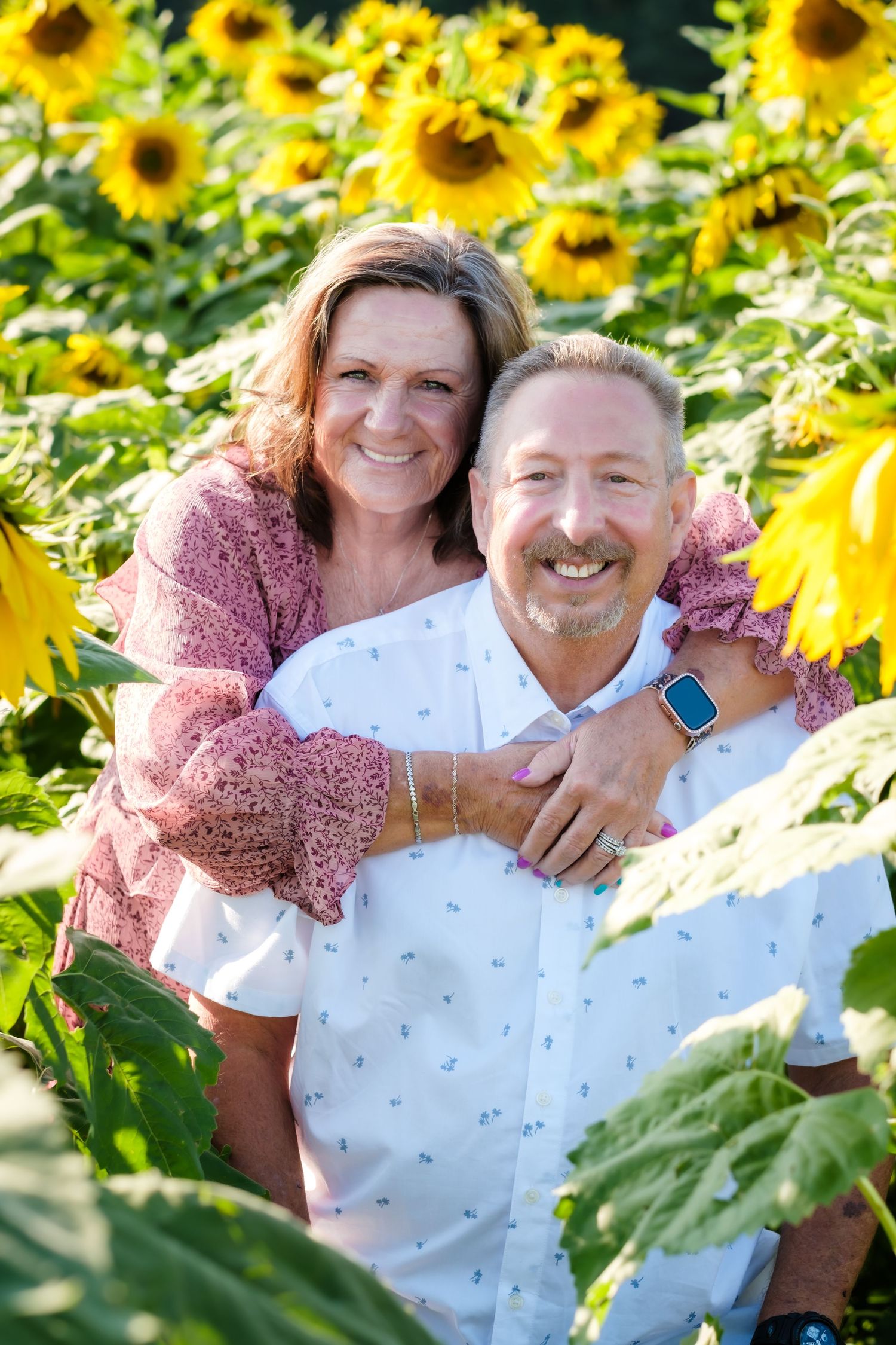 Smiling couple embracing in a vibrant sunflower field, woman in pink lace top hugging man in white shirt.