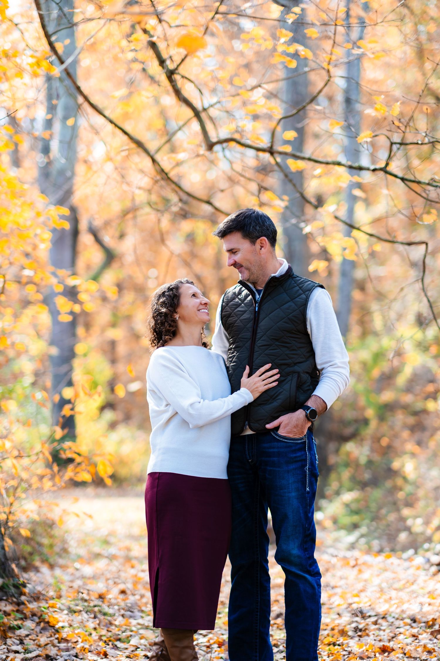 Couple smiling at each other in autumn forest, woman in white sweater and burgundy skirt, man in navy vest and jeans.