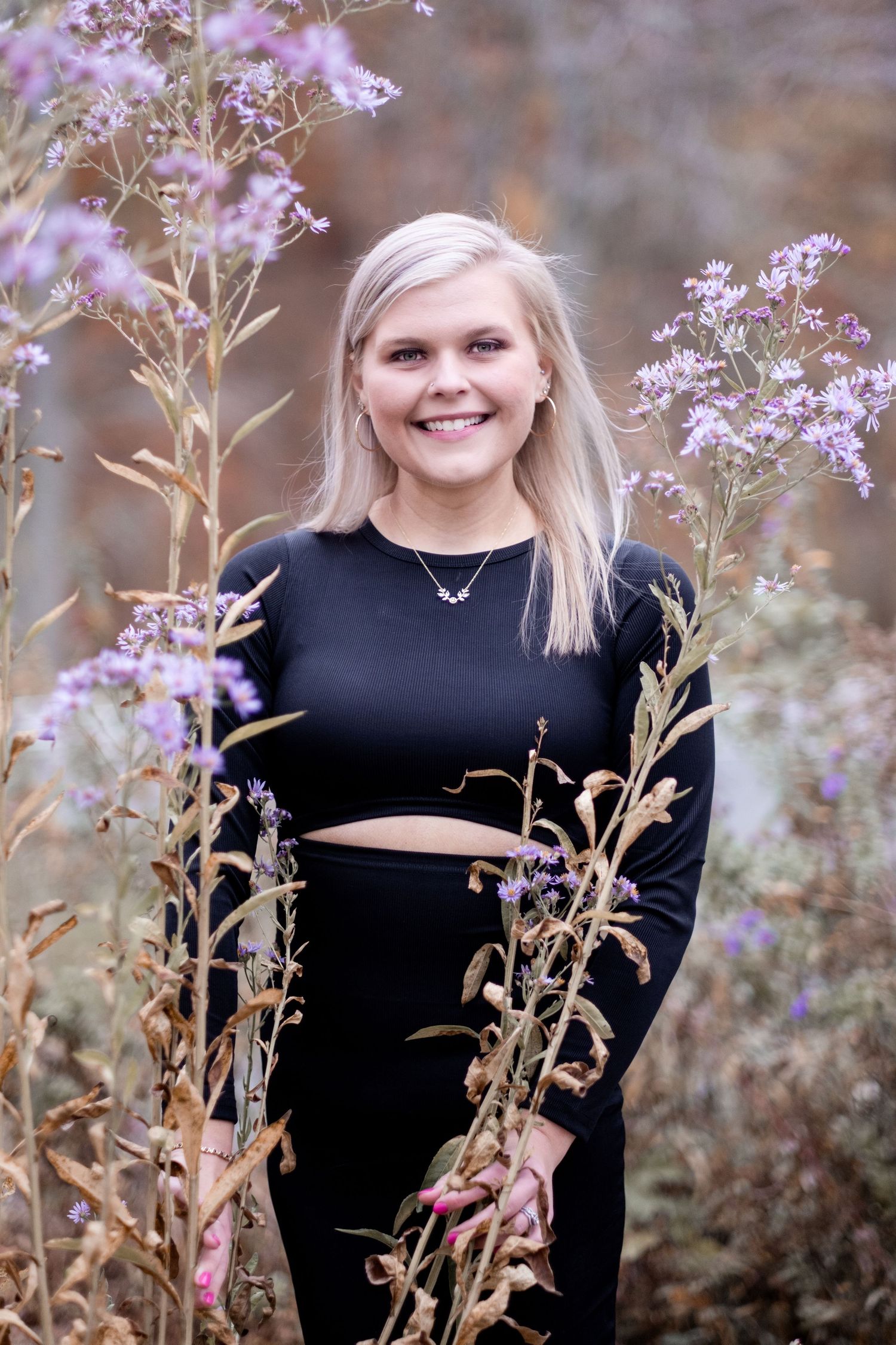 Smiling blonde woman in black crop top outfit standing among purple wildflowers in a natural outdoor setting.