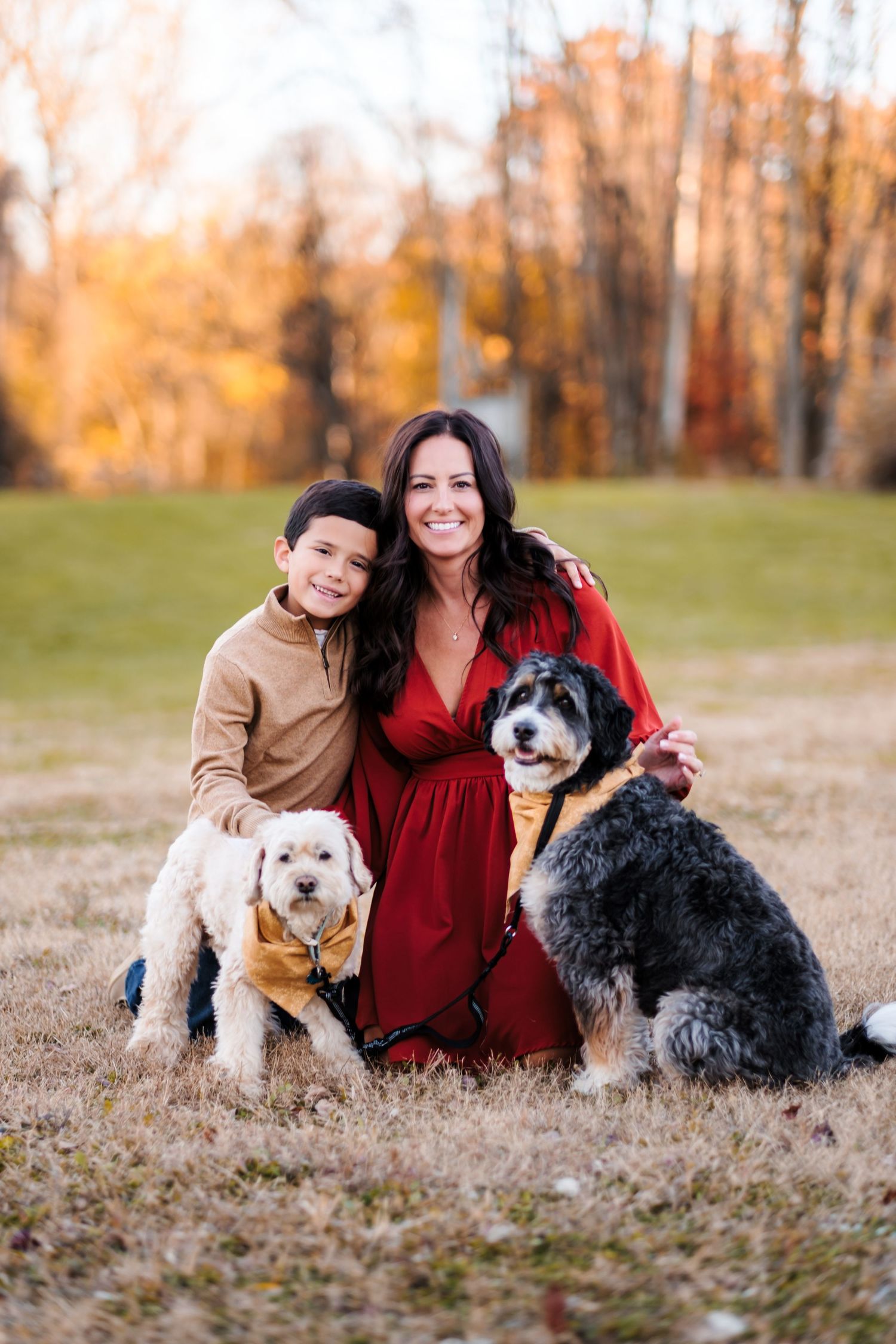 Woman in red dress poses with young boy and two dogs in a sunny autumn park setting.
