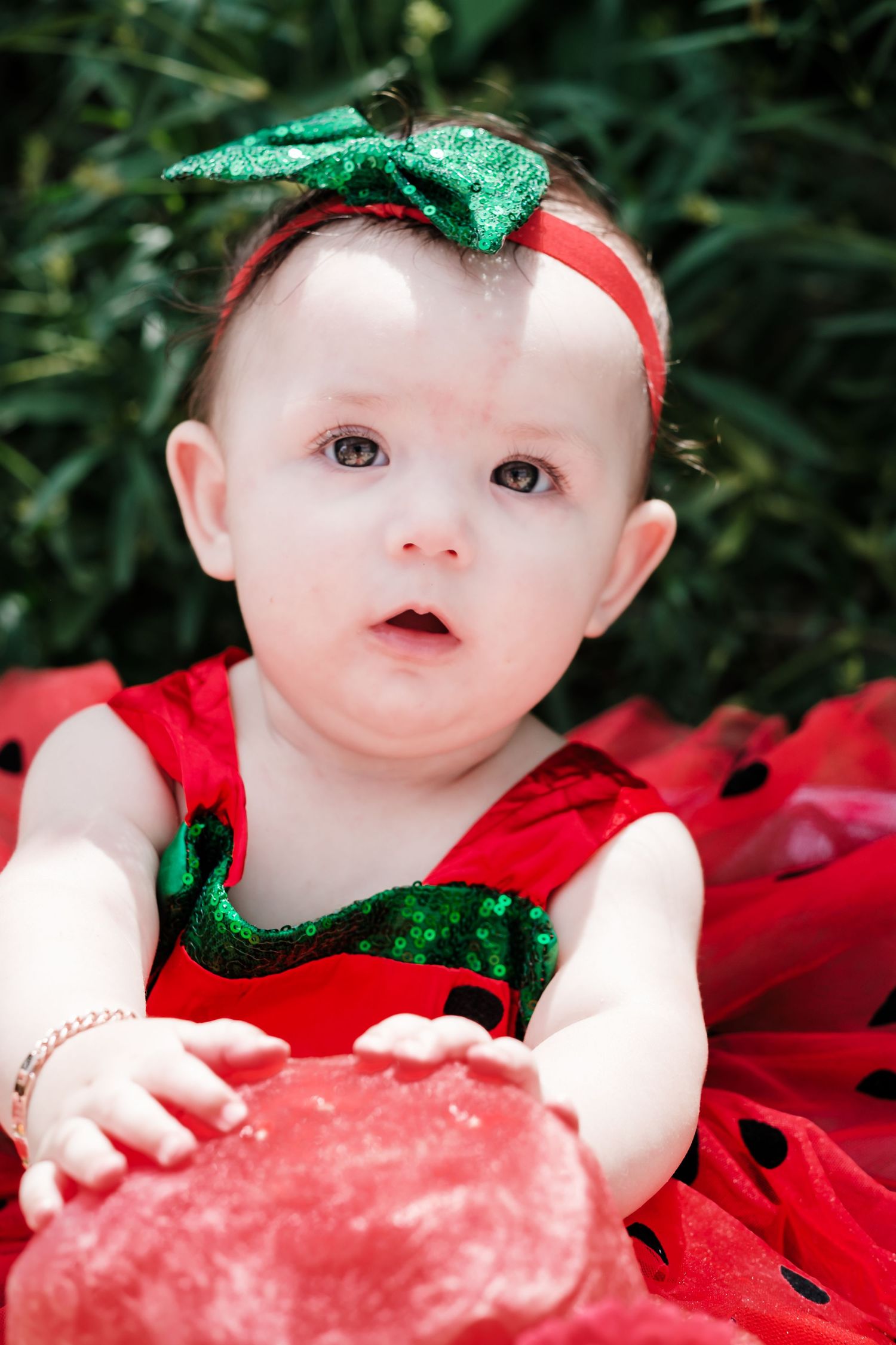 A cute baby girl in a red and green sequin dress with a green bow headband sitting outdoors in grass.