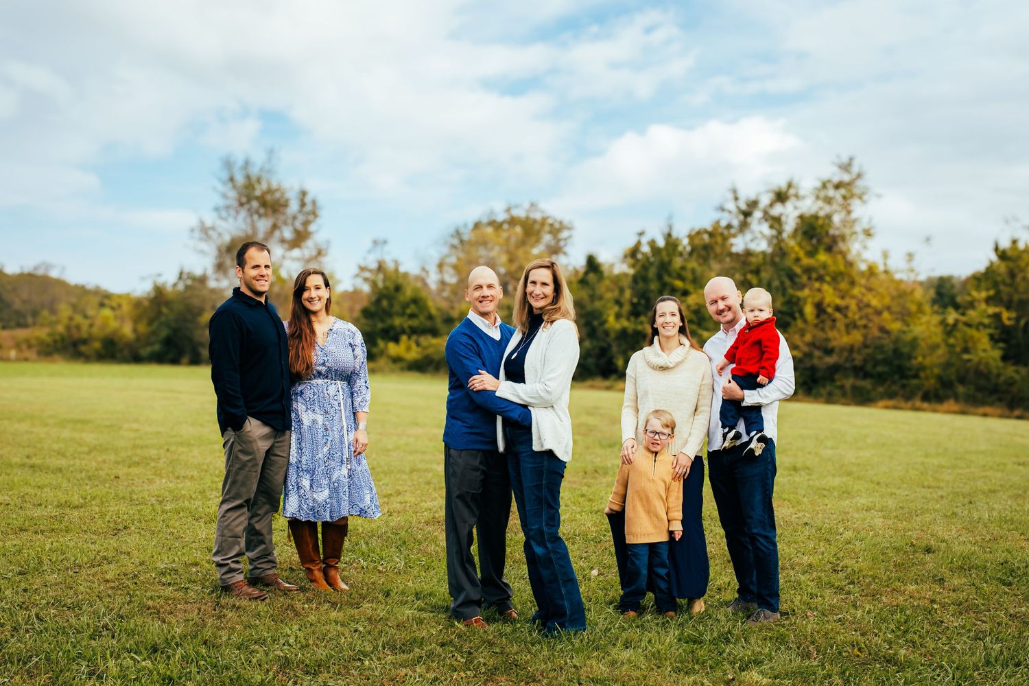 Family portrait of seven people posing outdoors in a green field with trees and cloudy sky in background.