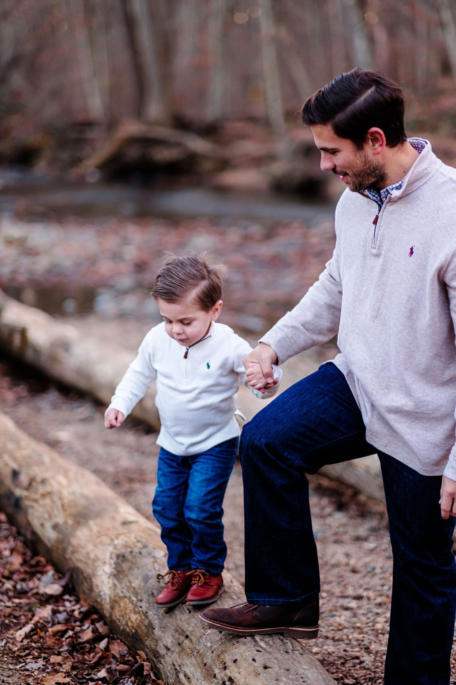 Father helps young son balance on a fallen log in a wooded area, both wearing matching Ralph Lauren sweaters.