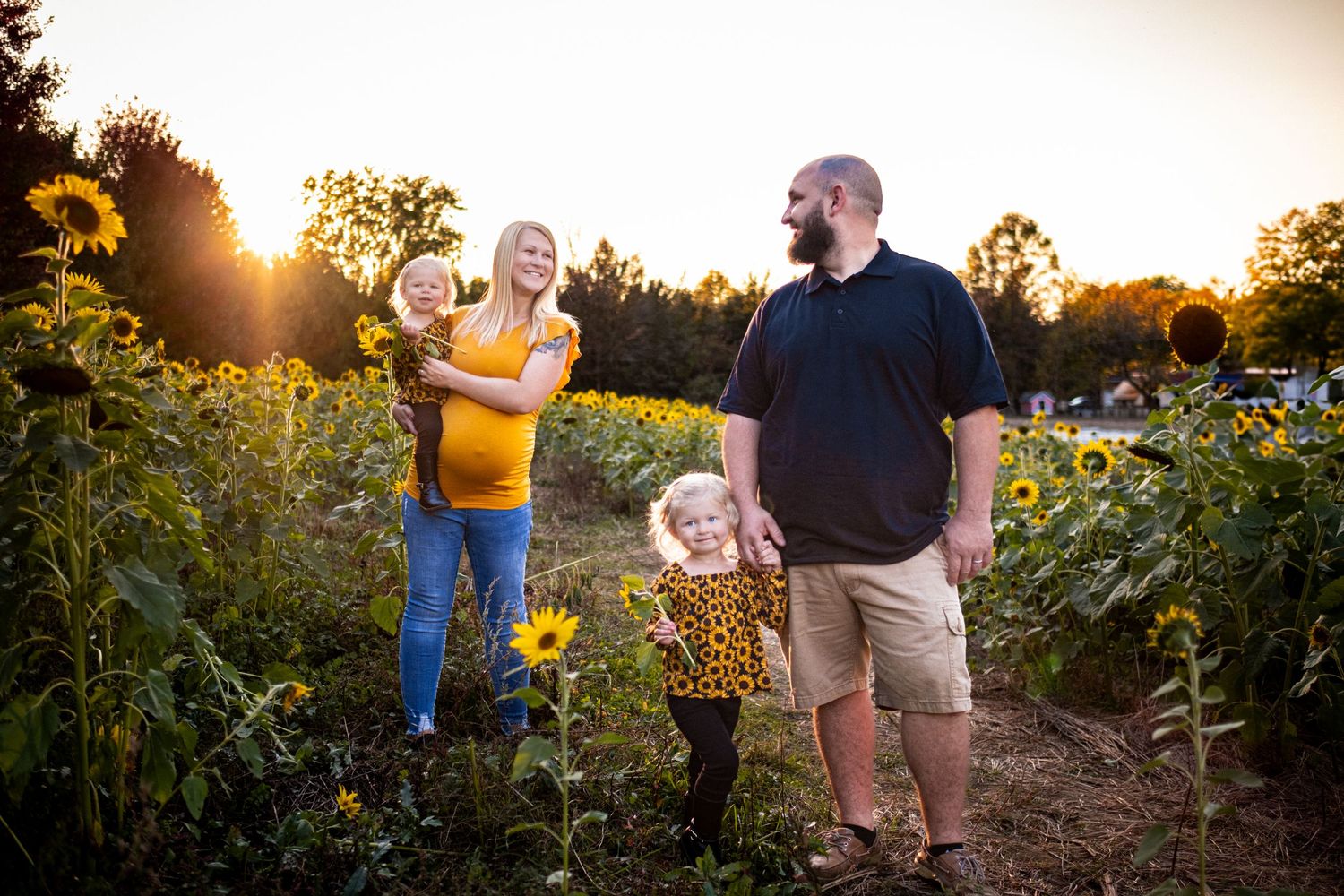 Family of four smiling in a sunflower field at golden hour sunset, two young girls with parents.