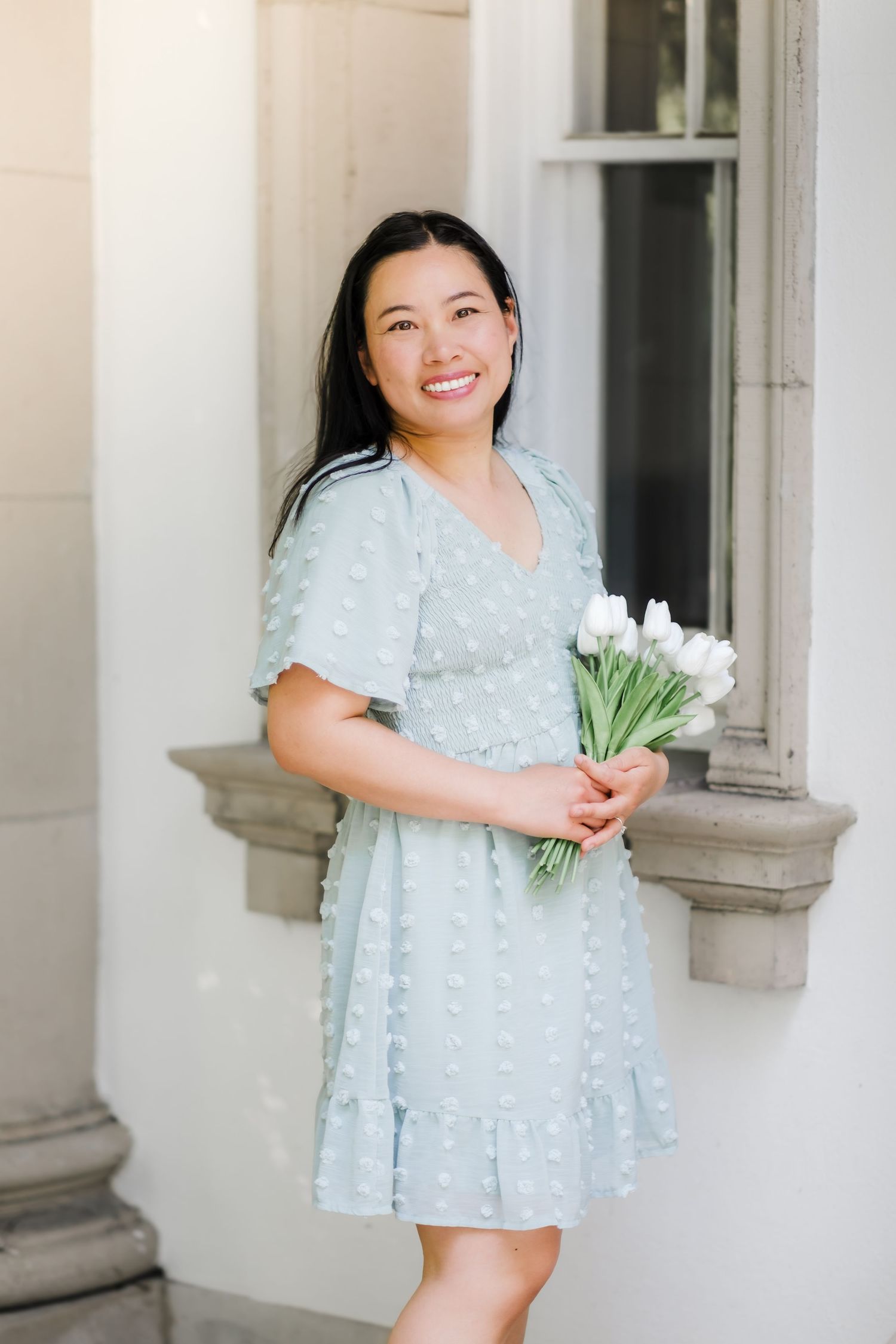 A person in a light blue dotted dress holds a bouquet of white tulips while standing by a stone pillar.