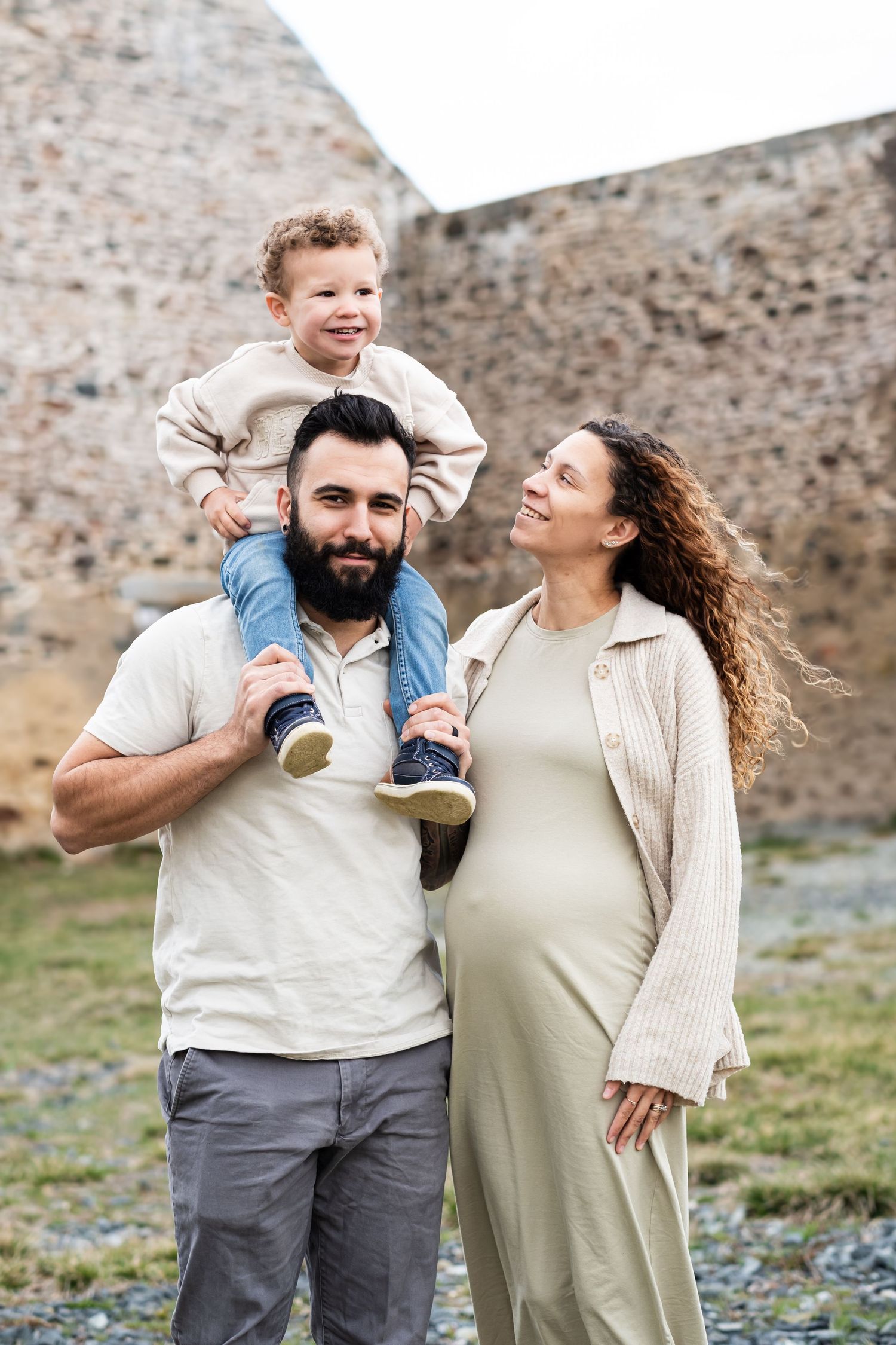 A happy pregnant woman and bearded man with a toddler on his shoulders pose outdoors near a stone wall.