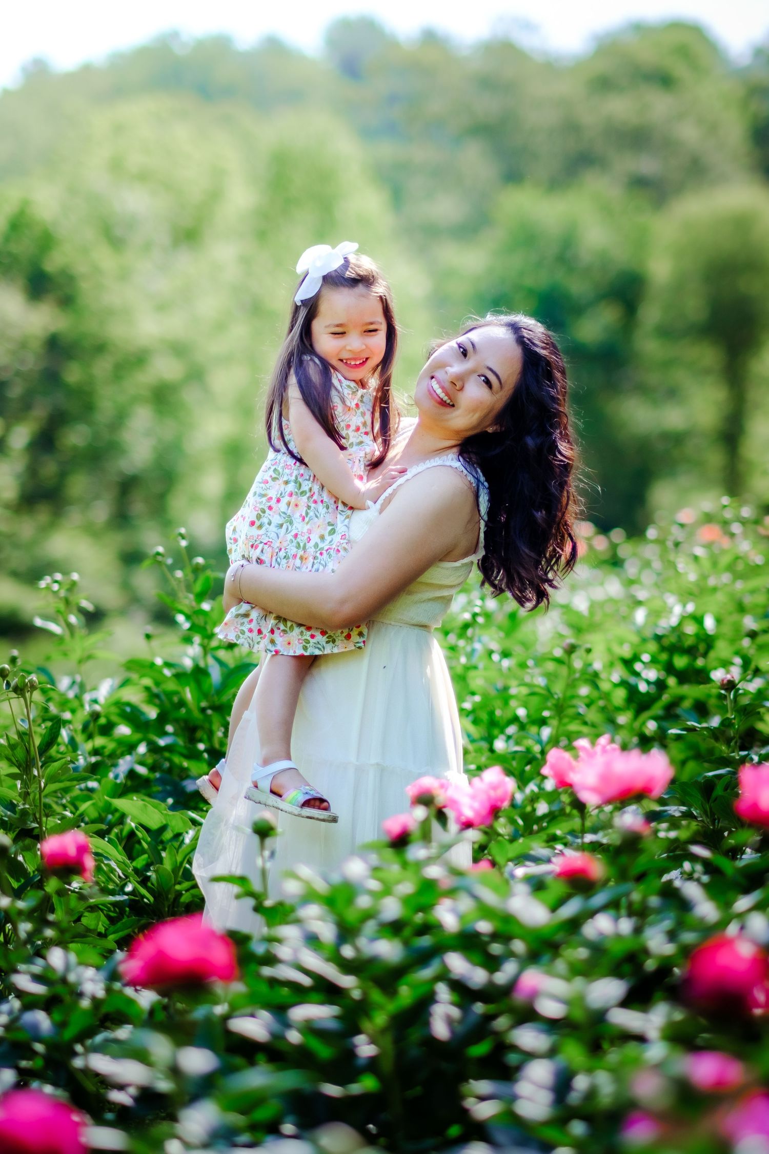 A smiling mother in a white dress holds her happy little girl in a floral outfit among pink peony blooms in a garden.