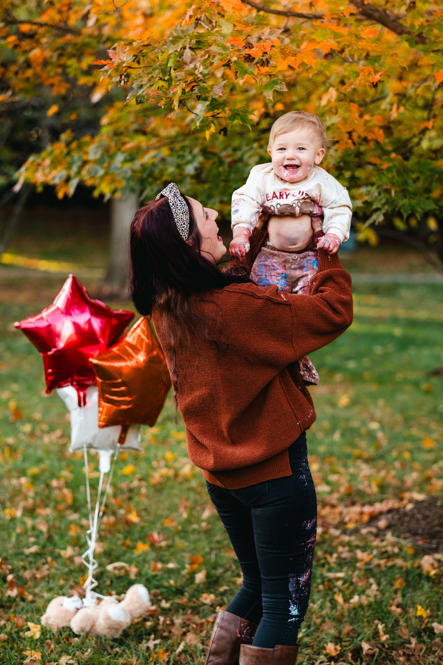 A mother lifts her laughing baby outdoors in autumn, holding red star balloons among colorful fall foliage.