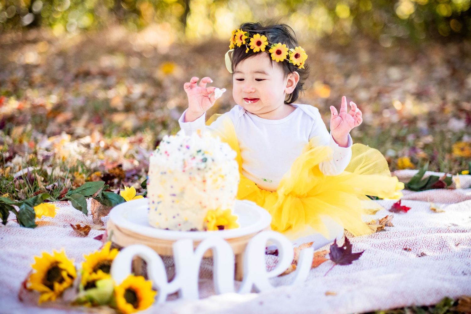 Baby girl in yellow tutu and floral crown celebrates first birthday with cake smash outdoors among sunflowers and autumn leaves.