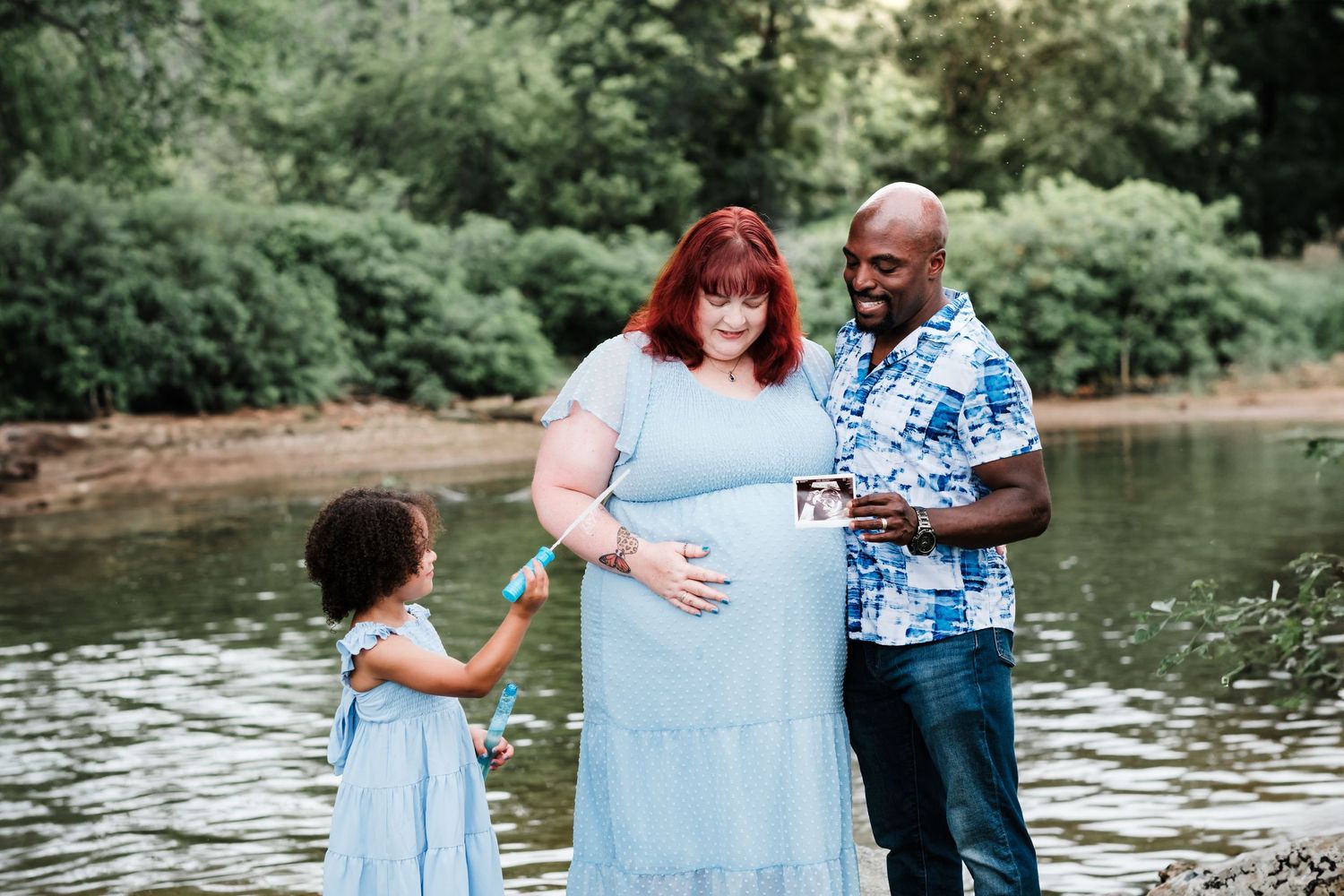 Pregnant woman in blue dress with husband and daughter by a river during a maternity photo session.