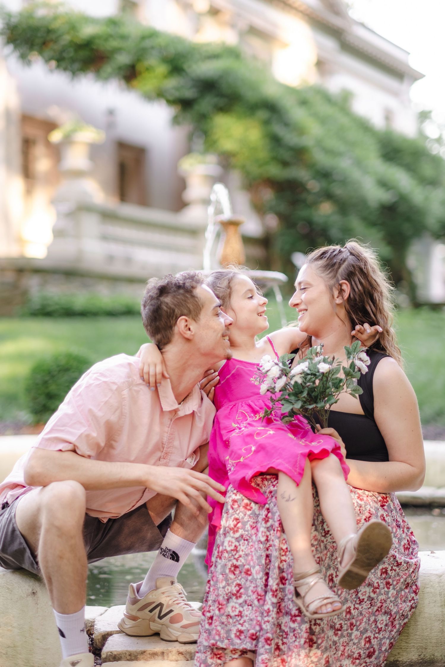 A happy family with a young girl in pink dress sharing kisses near a fountain outdoors.