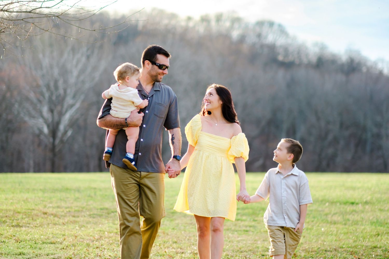 Happy family of four walking in a sunny field, parents holding hands with toddler and young boy.