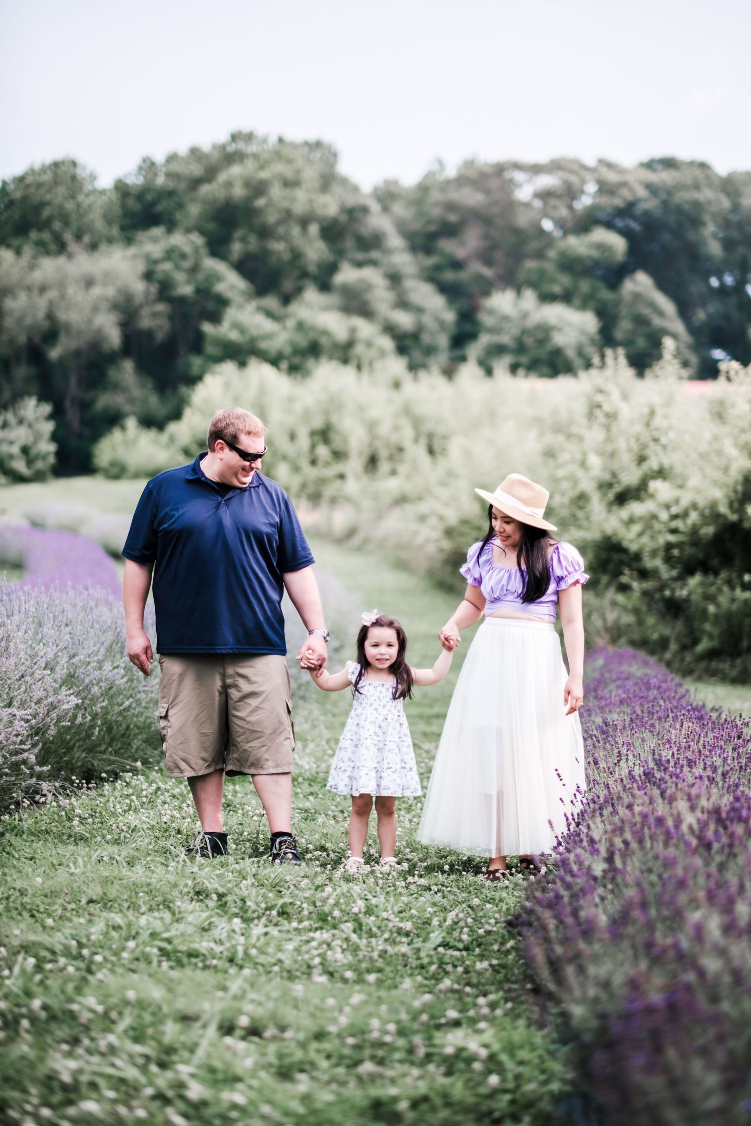 Family of three holding hands in a lavender field, parents smiling at young daughter in white dress on a summer day.