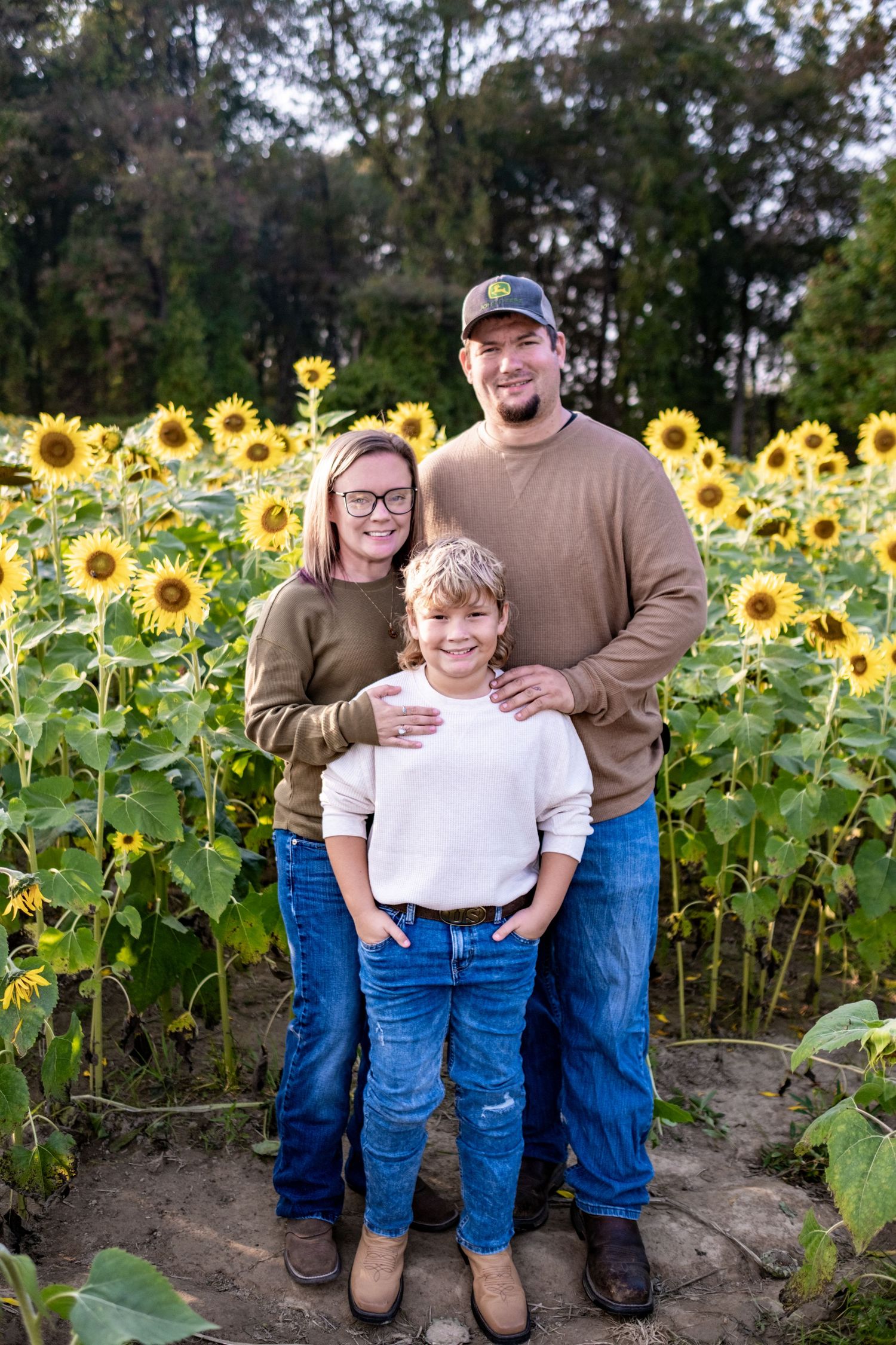 Family of three posing in a sunflower field, wearing casual fall outfits including jeans and earth-toned sweaters.