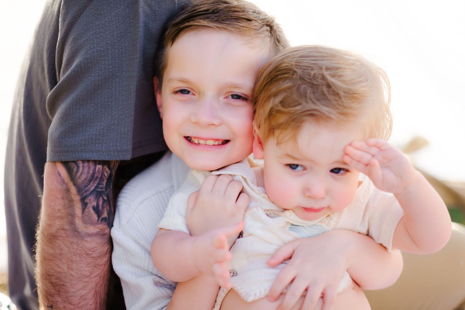 Older boy smiling and hugging a baby sibling, with an adult standing behind them in a warm outdoor setting.