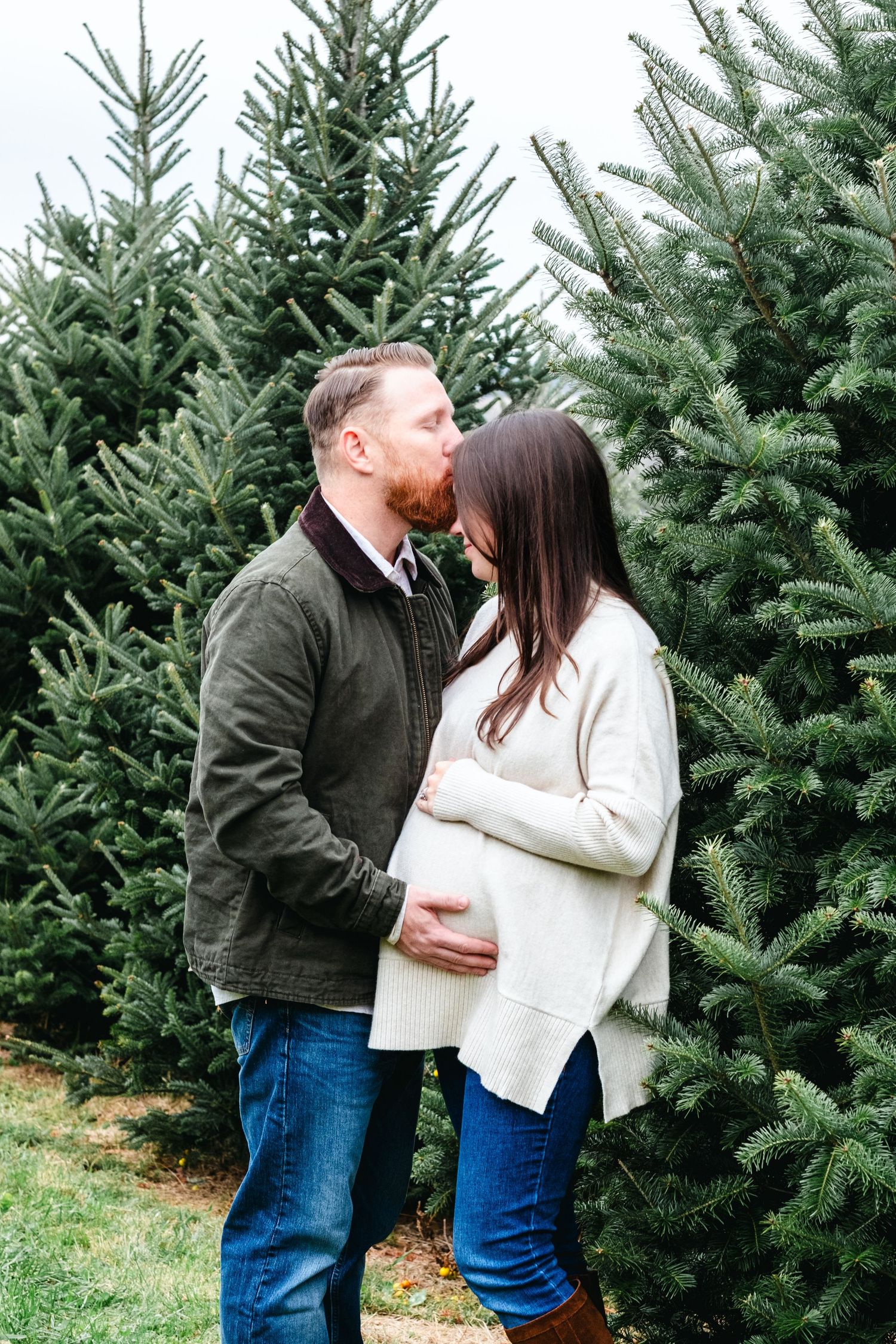 Couple sharing a tender kiss at a Christmas tree farm, woman in cream sweater cradling baby bump.