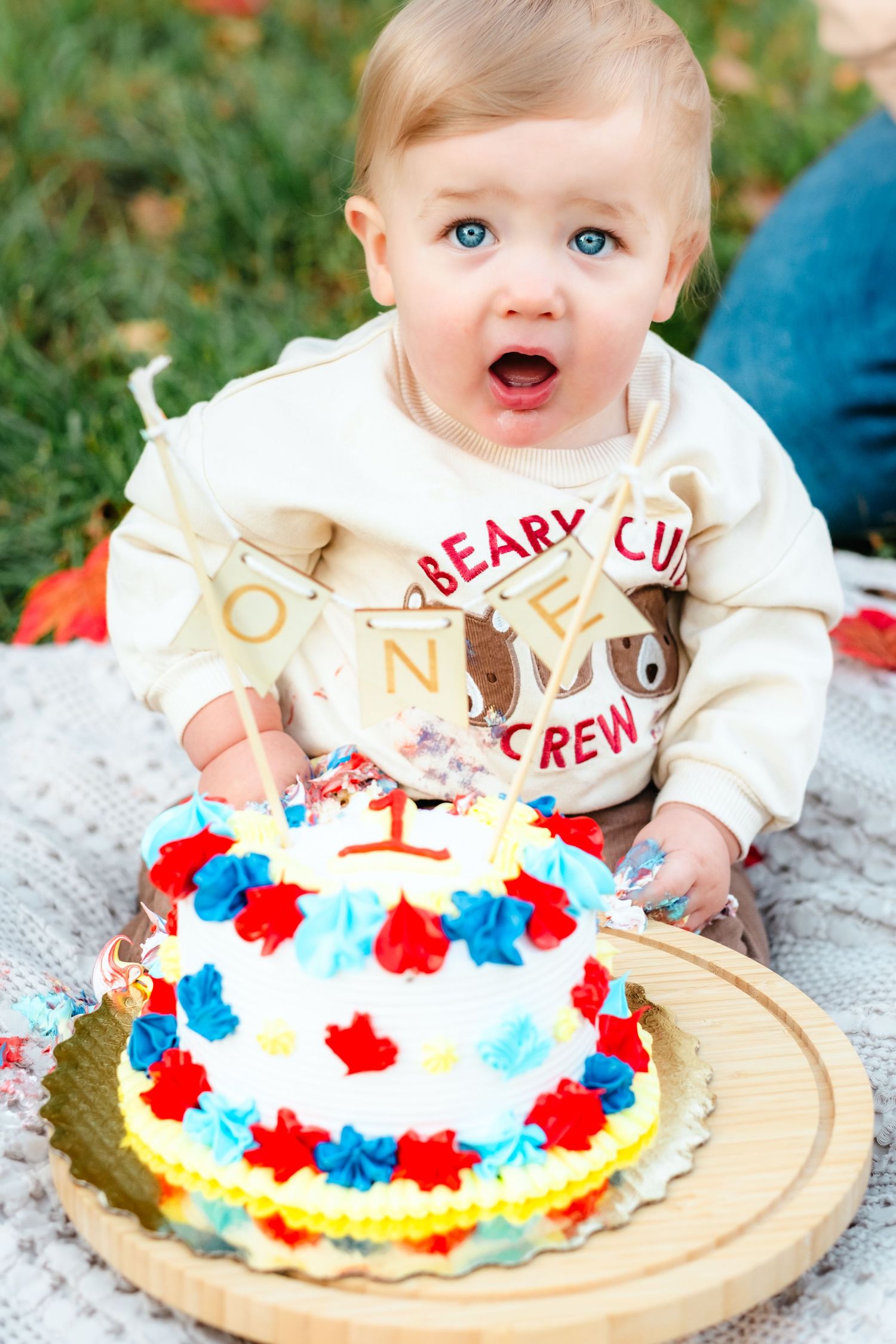 Blue-eyed baby celebrating first birthday with colorful cake outdoors, wearing a Beary Cute Crew sweatshirt.