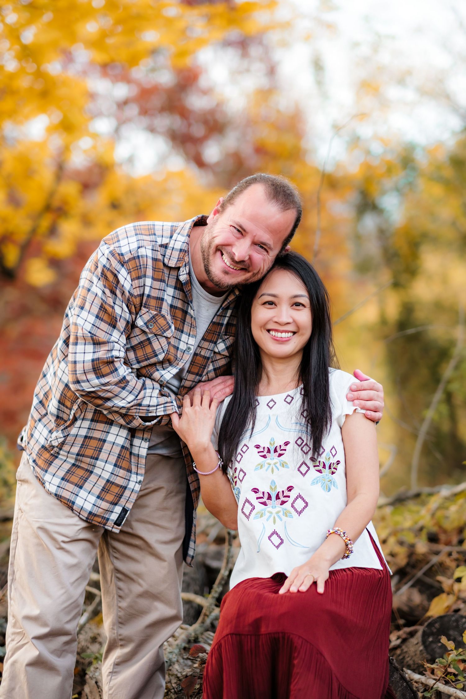 Couple smiling together outdoors in autumn, man in plaid shirt embracing woman in white embroidered top and red skirt.