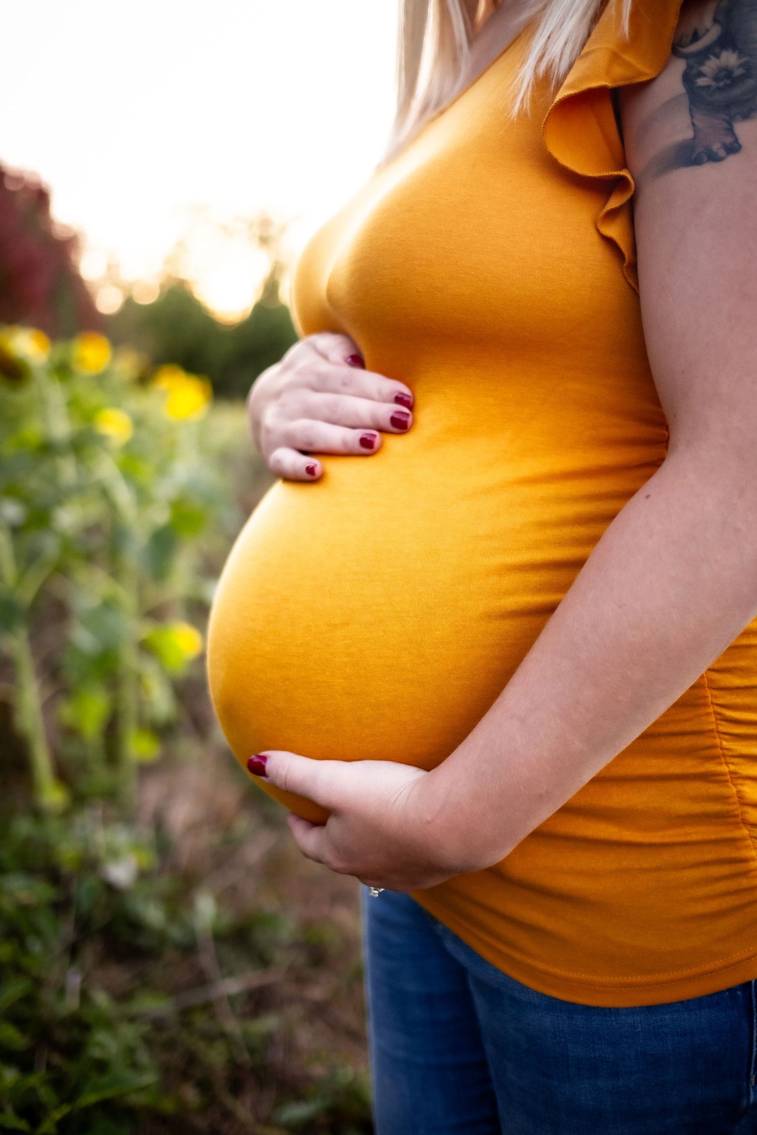Pregnant woman in orange top cradling baby bump outdoors in sunflower field at golden hour.