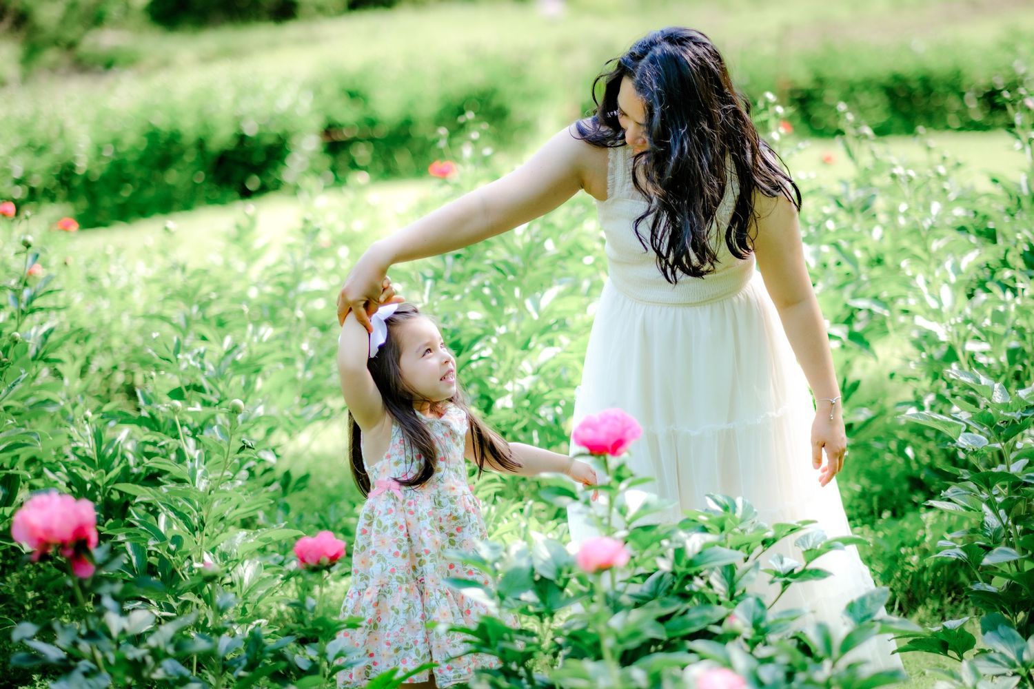 Mother in white dress twirling young daughter in floral dress among pink peonies in lush garden.
