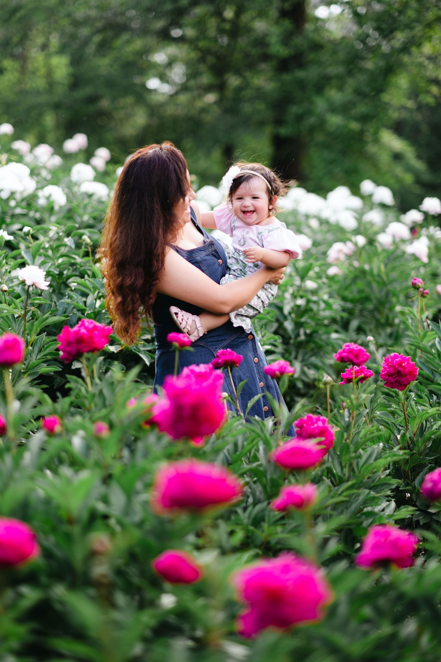 Mother holding laughing baby girl among vibrant pink and white peony flowers in a lush garden.